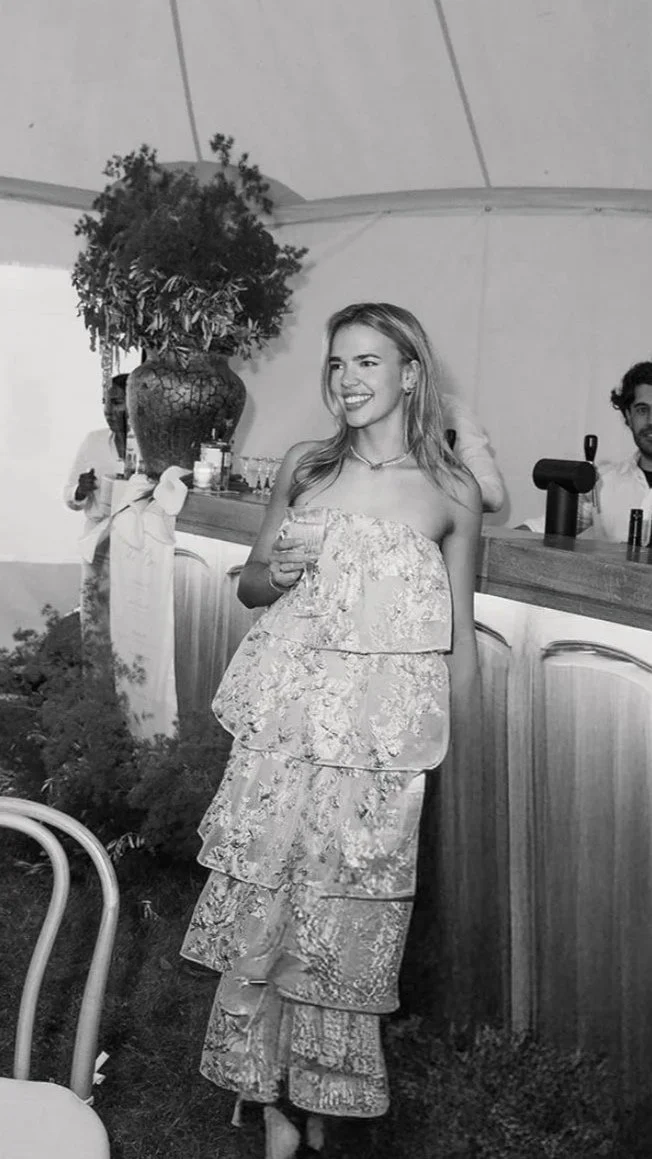 A woman in a strapless, layered floral dress holding a glass of wine at a social event or party, standing near a bar with a large vase and people in the background.