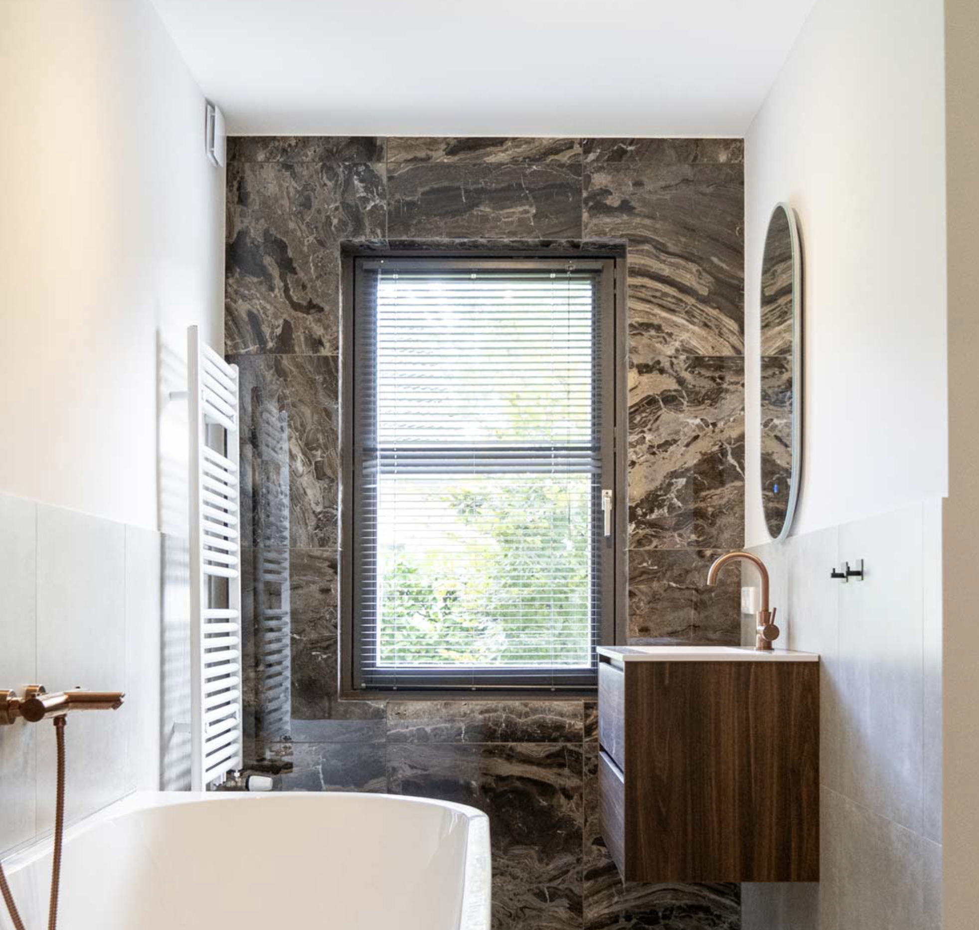 Modern bathroom with large window, marble wall tiles, a white bathtub, wall-mounted wooden vanity with copper faucet, oval mirror, and white towel warmer.