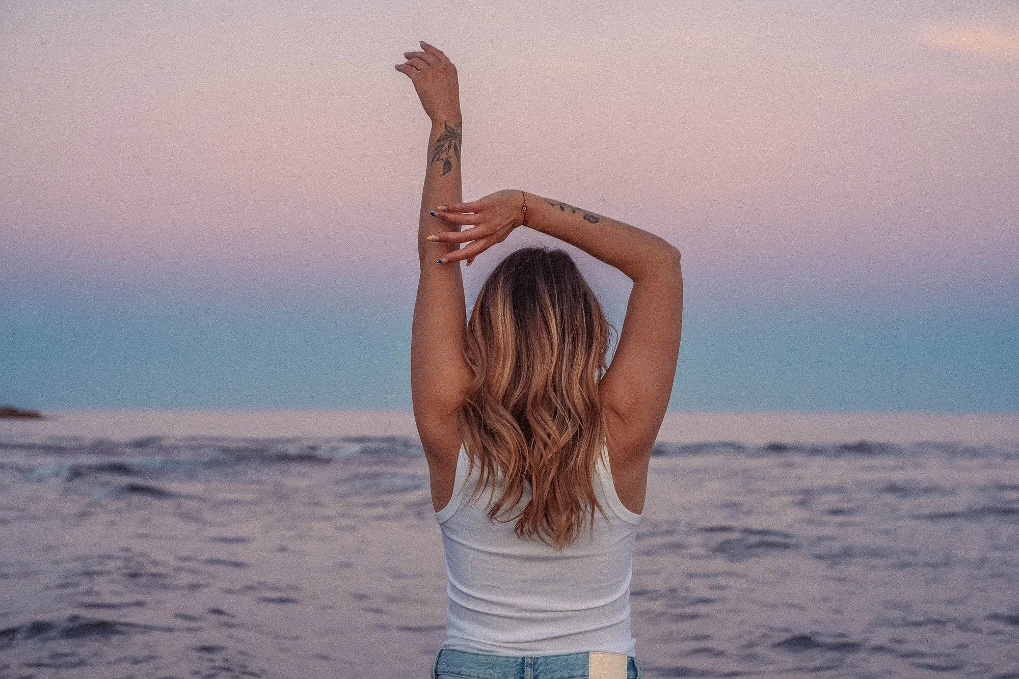 Une femme de dos, avec des tatouages sur les bras, se tient sur la plage au coucher du soleil, levant les bras vers le ciel.