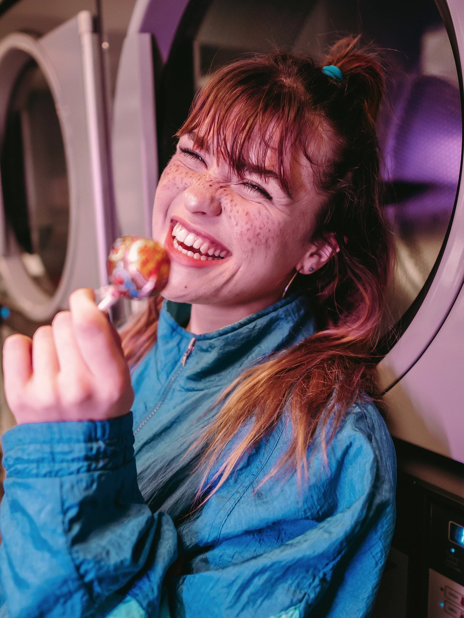 Jeune femme souriante tenant une sucette en main, portant une veste bleue, dans une laverie automatique.