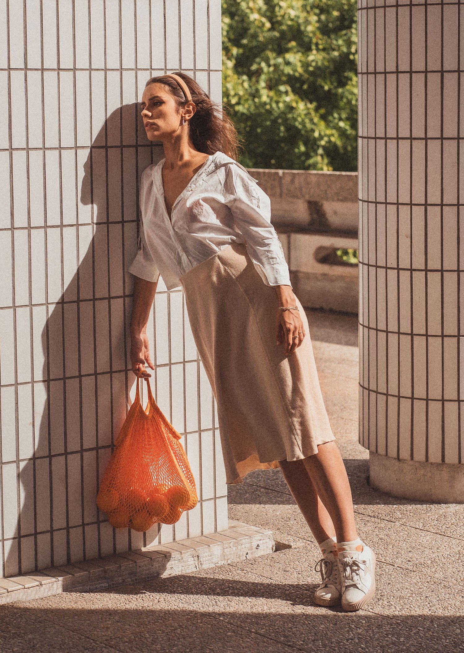 Une femme portant une robe beige et un Chemisier blanc, tenant un sac orange en filet, se tient contre un mur en céramique, avec des arbres verts en arrière-plan, sous une lumière du soleil.