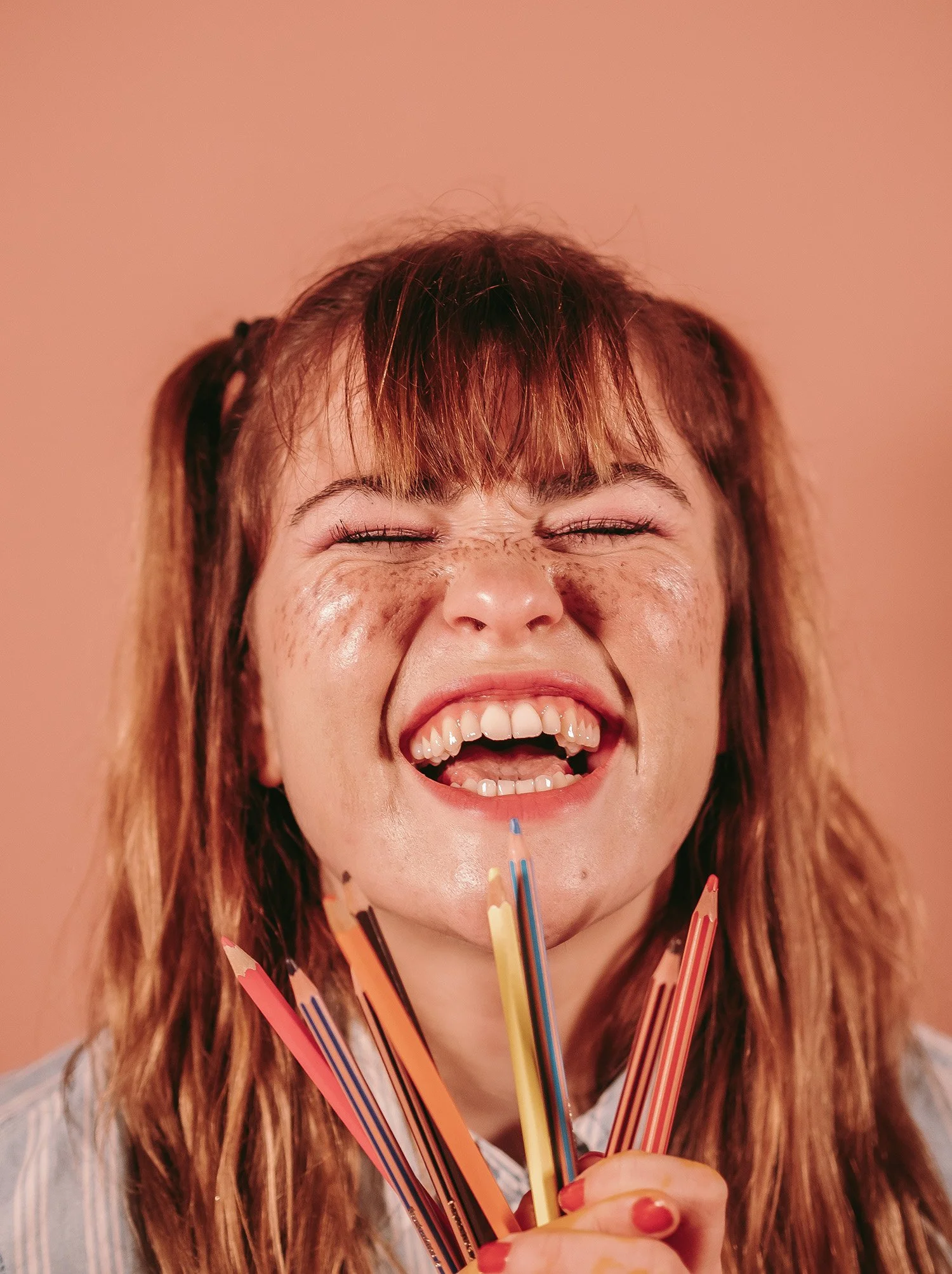 Une femme souriante avec des cheveux longs et roux, tenant plusieurs crayons de couleur devant son visage, avec un mur beige en arrière-plan.