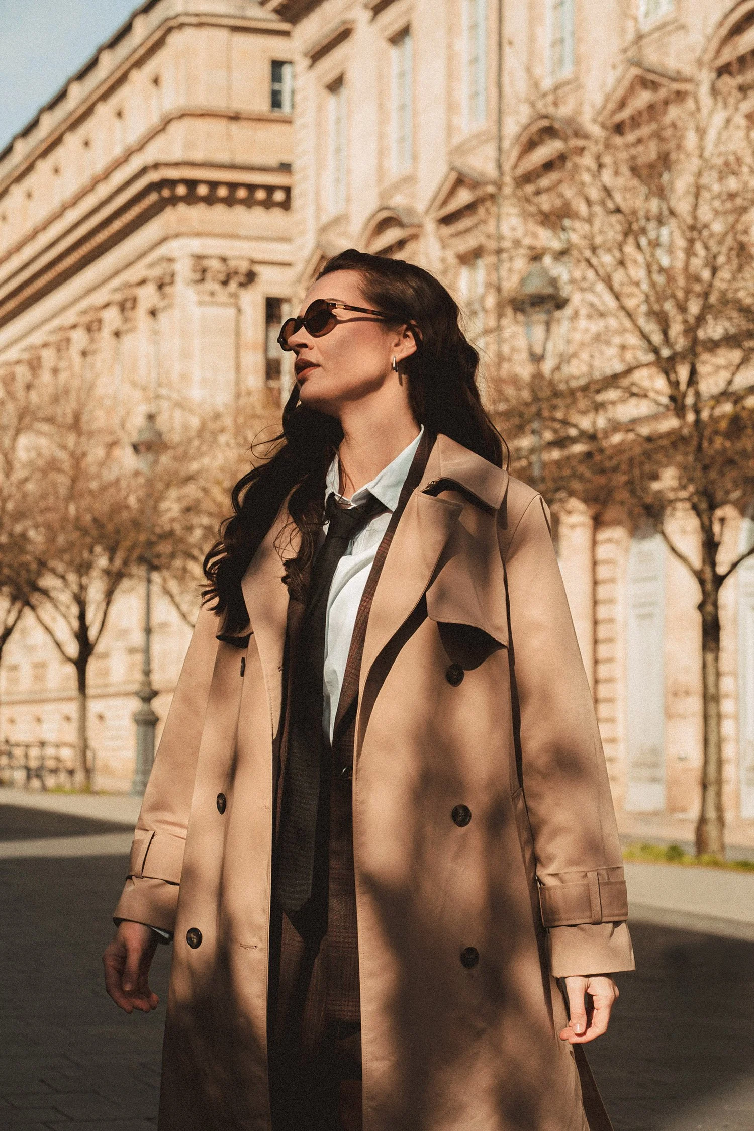 Une femme portant un trench beige, des lunettes de soleil foncées, une chemise blanche et une cravate noire, marche dans une rue avec des bâtiments anciens en arrière-plan, à l'automne.