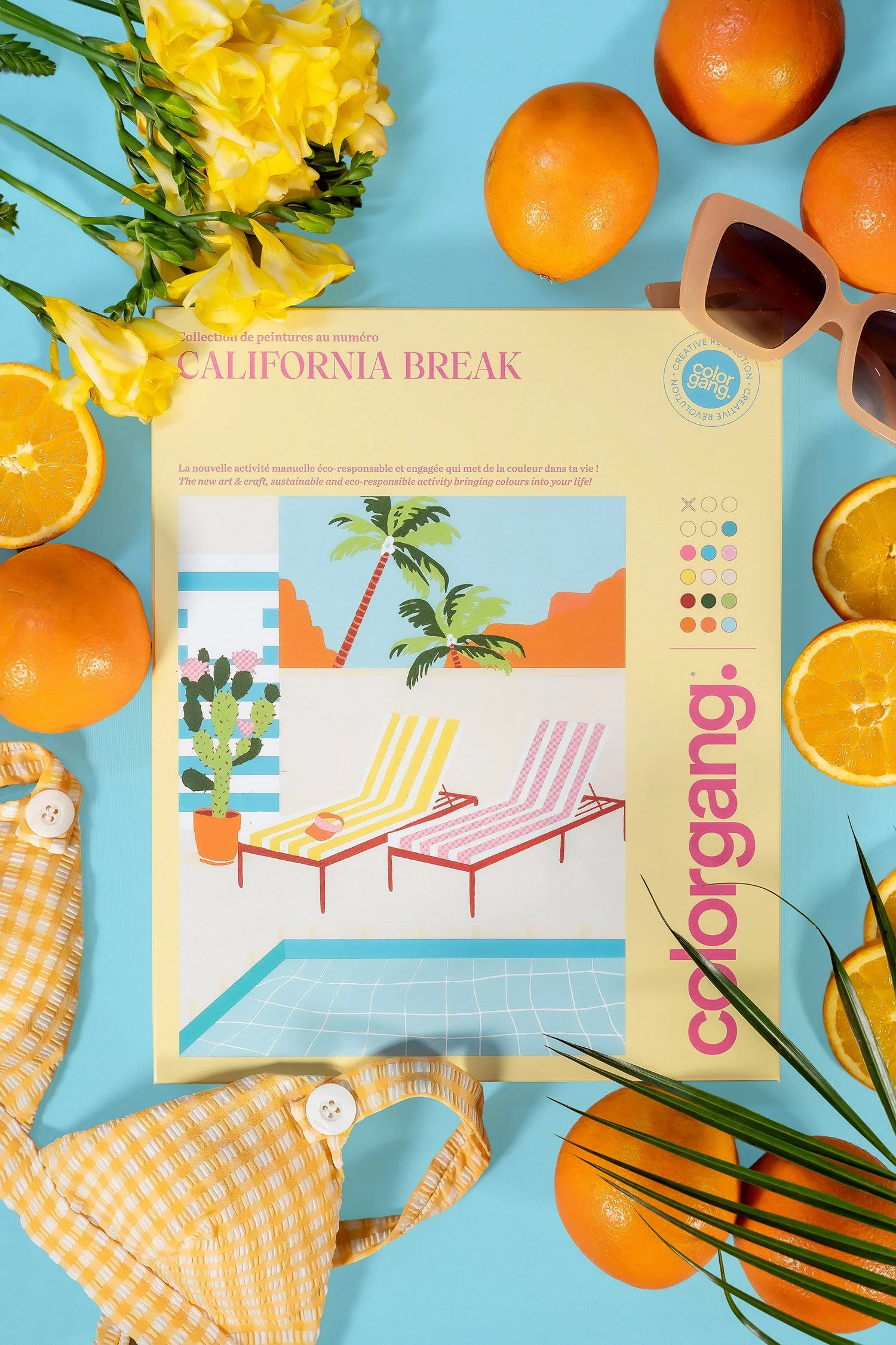 Une mise en scène de fruits oranges, des lunettes de soleil beige, un livret de peinture avec un thème californien, un peignoir jaune à carreaux, une palme, et des boutons blancs sur une surface bleu clair.