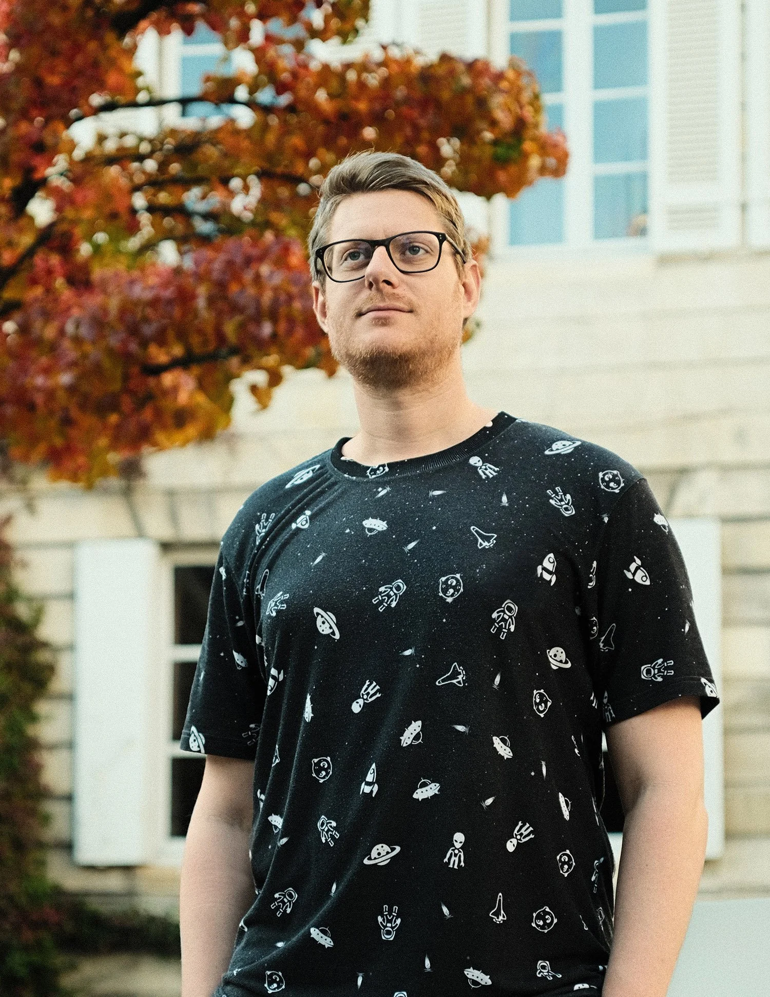 Jeune homme portant un t-shirt noir avec des motifs d'astronautes, de planètes et de fusées, portant des lunettes, dans un environnement extérieur avec des feuilles d'automne en arrière-plan.