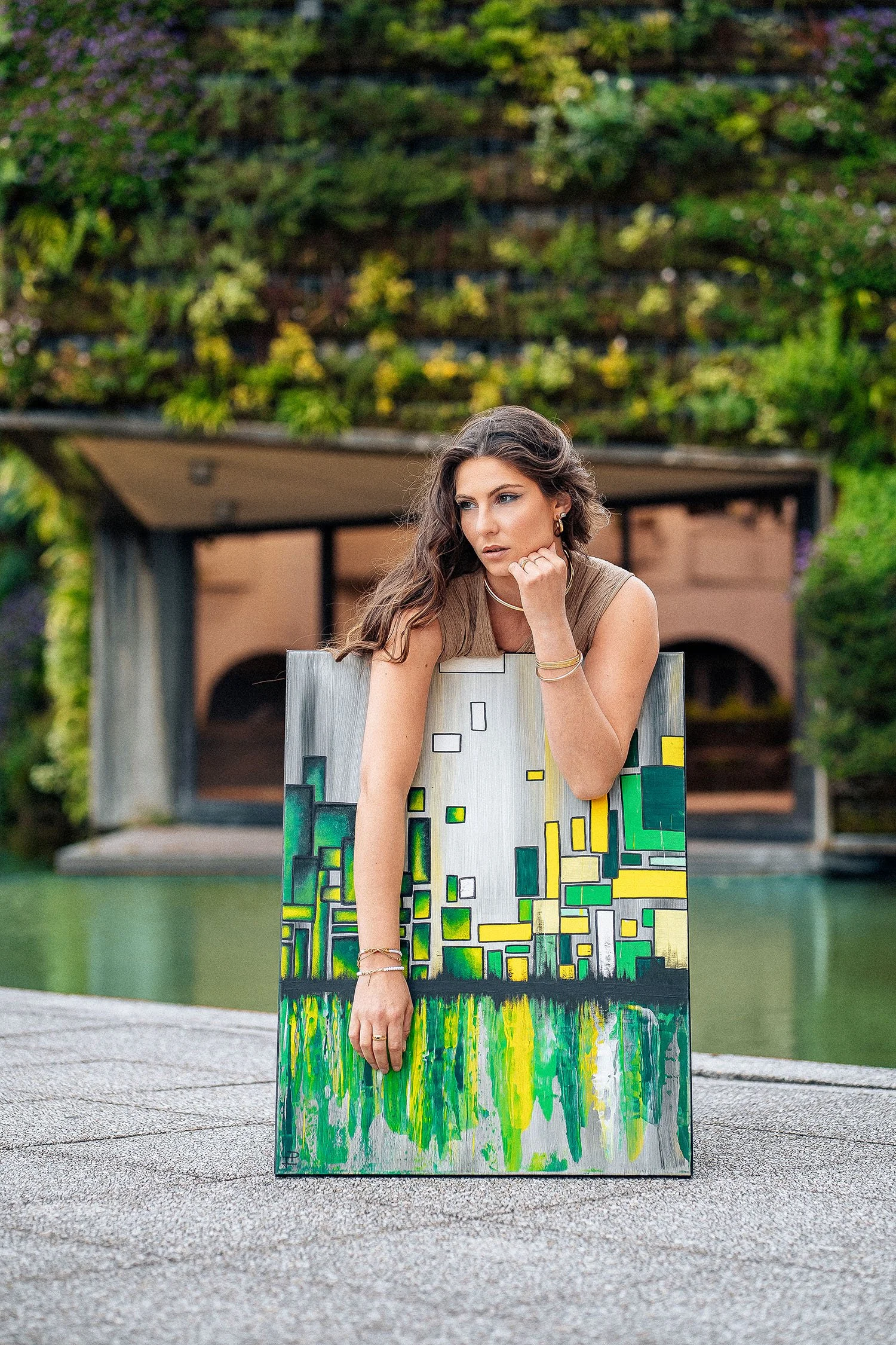 Femme posant derrière une œuvre d'art représentant une ville, en extérieur avec un jardin vert en arrière-plan.
