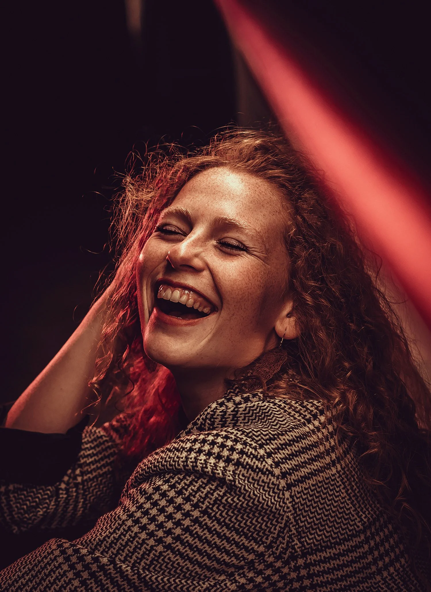 Une femme souriante avec des cheveux bouclés roux, portant un vêtement à carreaux, dans un décor obscur avec un éclairage chaud.