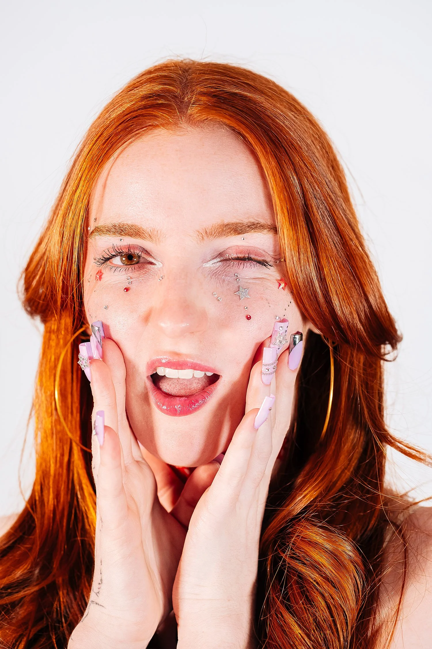 Une femme aux cheveux roux, portant des décorations de visage, fait un sourire expressif avec une expression de surprise ou de joie.