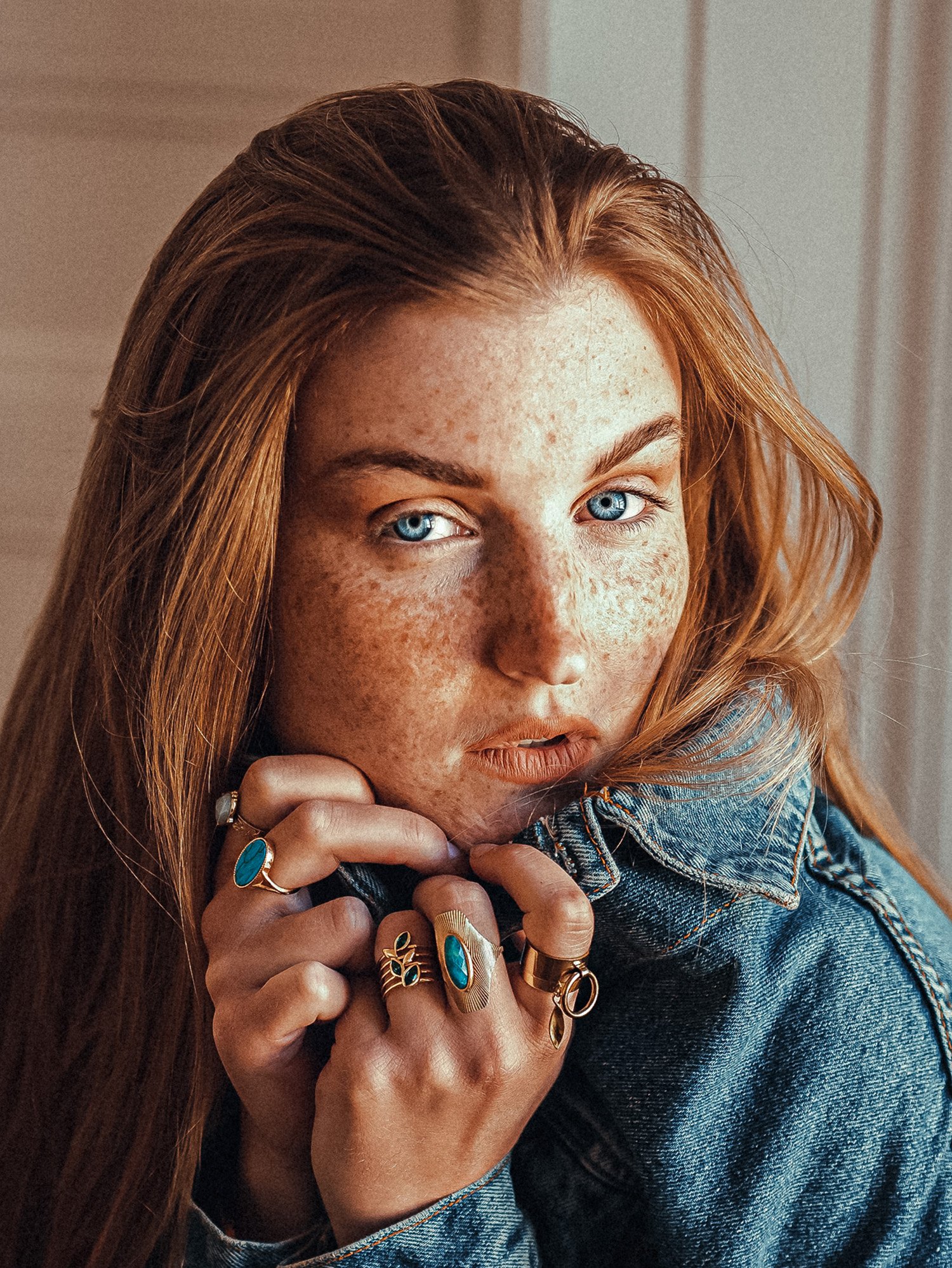 Un gros plan d'une femme avec des cheveux roux, des taches de rousseur, portant plusieurs bijoux en or avec des pierres turquoise, tenant sa main près de son visage.