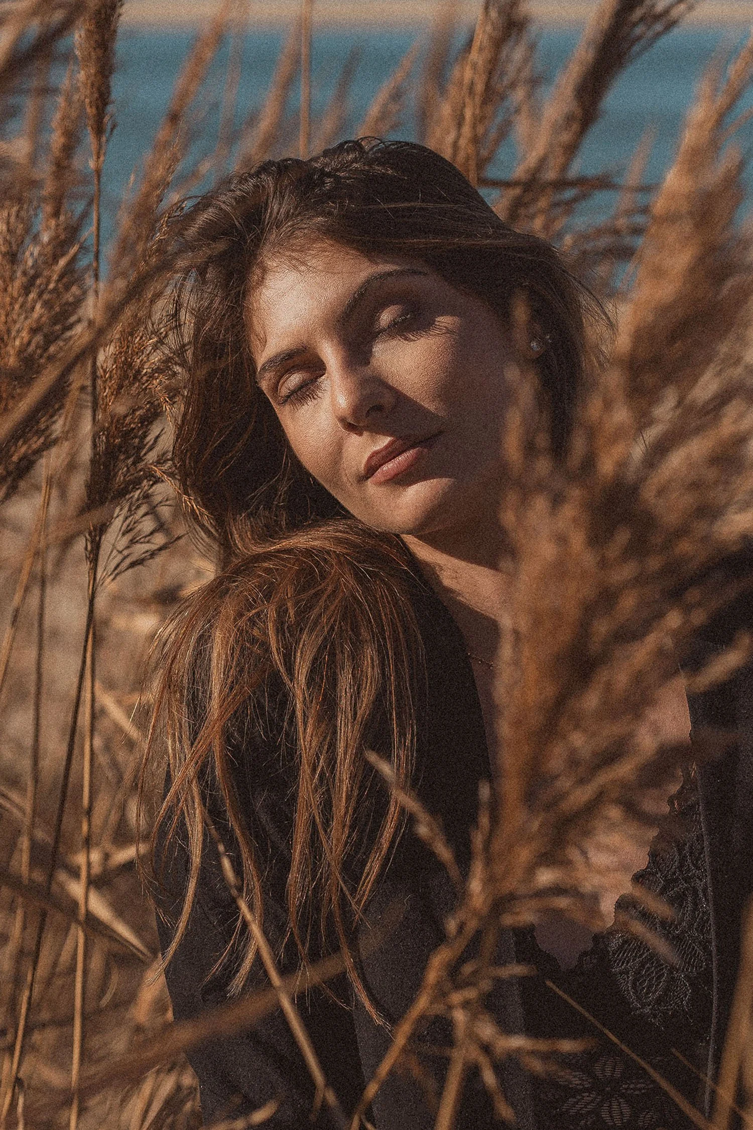 Une femme avec des cheveux longs et bouclés, yeux fermés, posée dans un champ de fourrage sec et de couleurs chaudes, en plein soleil.