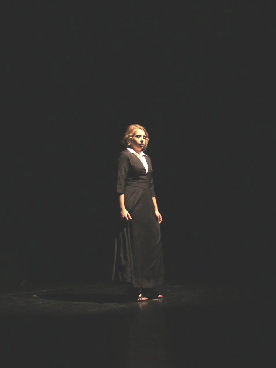 Mujer con vestido largo negro en un escenario oscuro, iluminada por una luz centrada en ella.