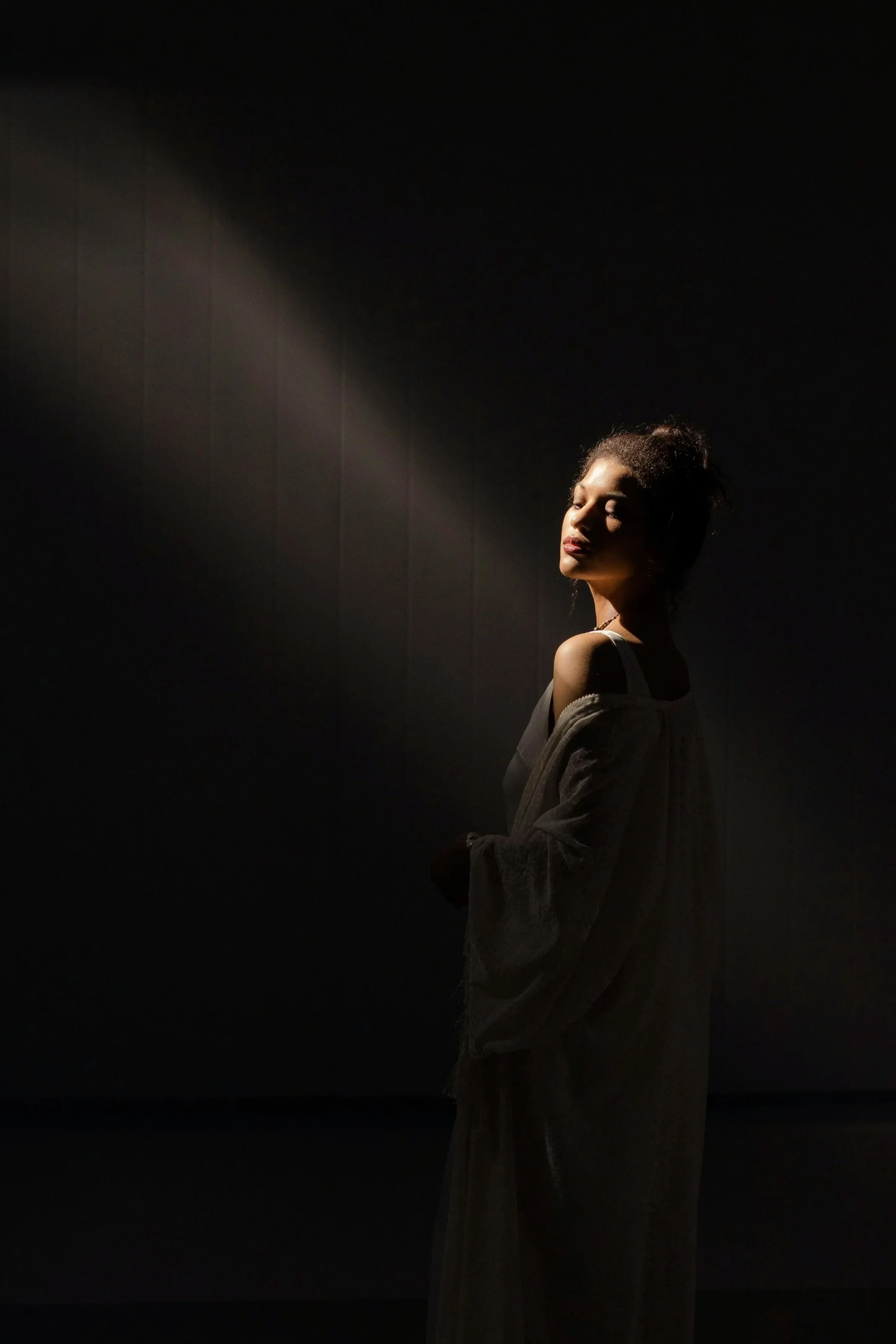 Mujer en la oscuridad, iluminada por un haz de luz, con expresión contemplativa y vestido suelto, en un ambiente sombrío.