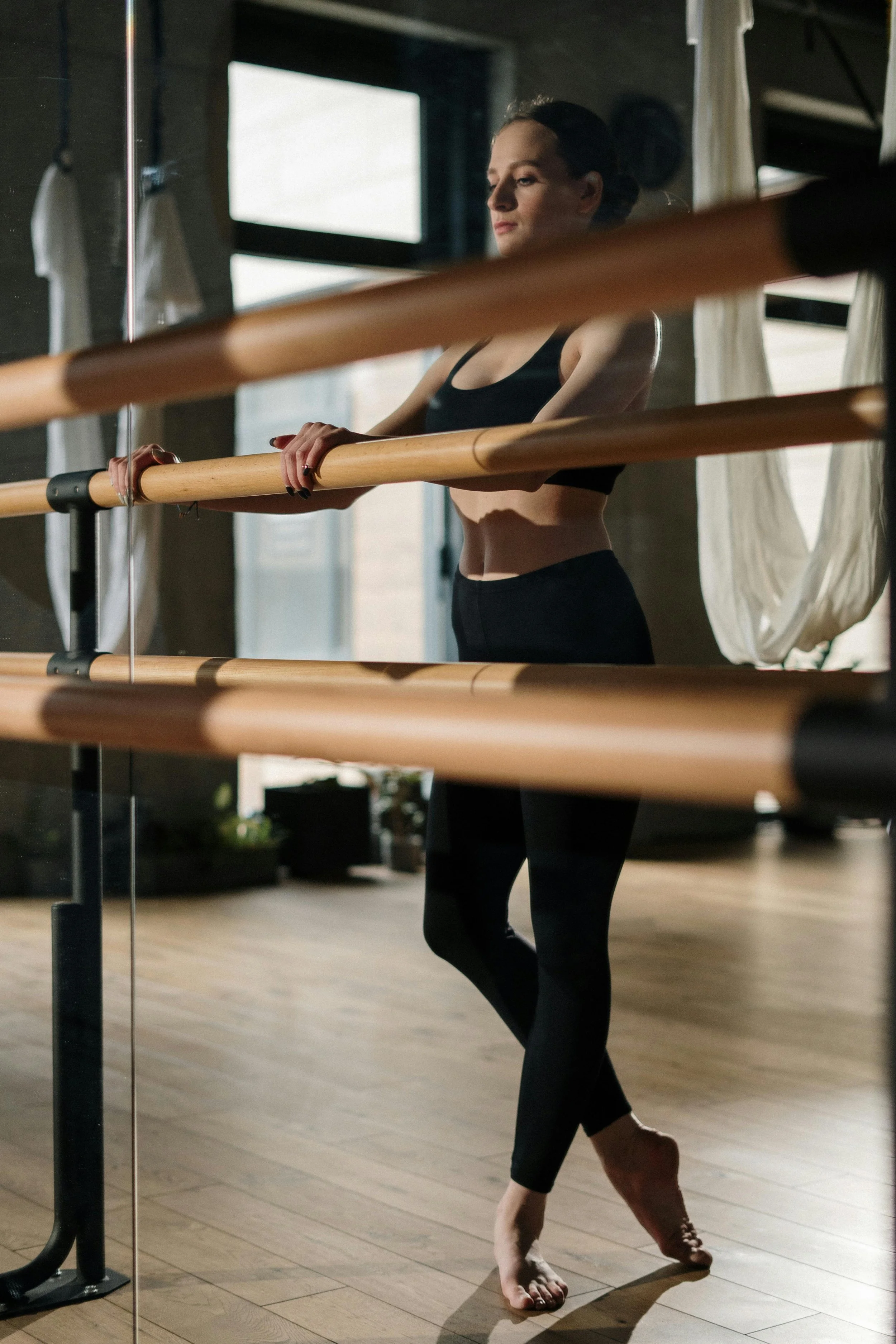 Mujer en ropa de ejercicio en un estudio de ballet, colocando las manos en una barra de ballet mientras practica, en un ambiente con ventanas grandes y pisos de madera.