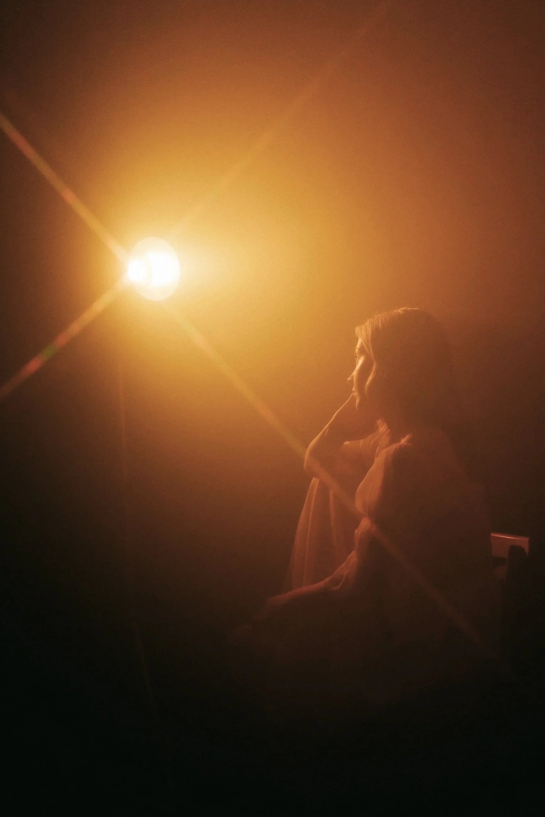 Mujer sentada en la oscuridad con una luz brillante al fondo, con cabello rubio y expresión pensativa.