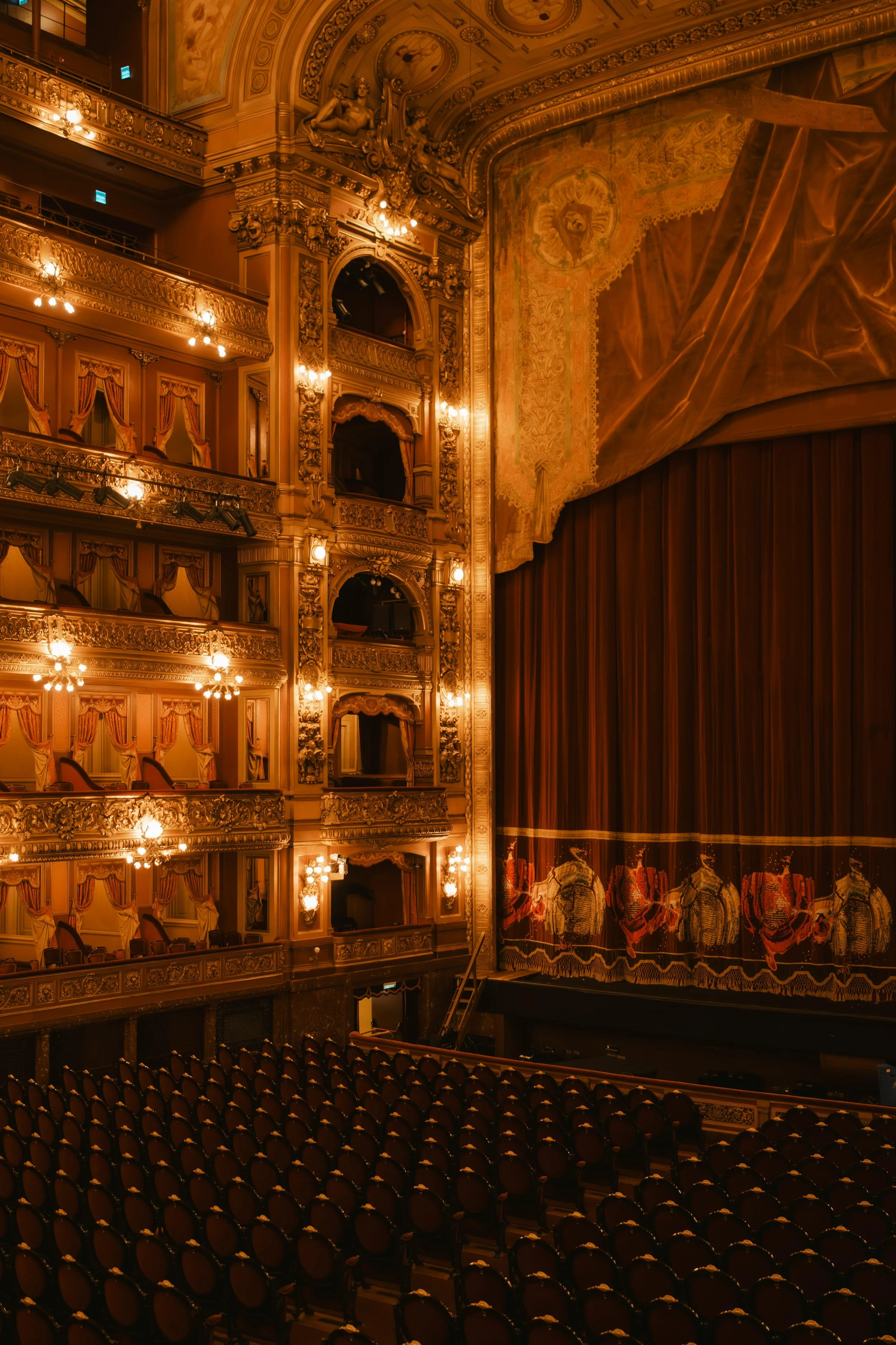 Interior de un teatro elegante con múltiples palcos y un escenario cubierto por cortinas rojas, decorado con detalles dorados y un telón con imágenes de caballos.
