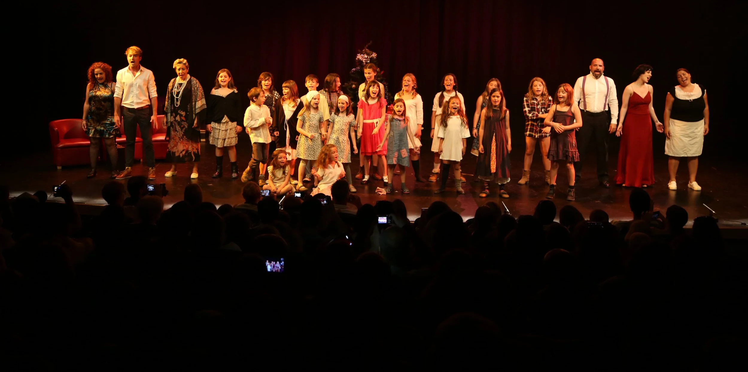 Grupo de niños y adultos en un escenario de teatro, algunos sonriendo y otros aplaudiendo, con un árbol de Navidad decorado en el fondo.