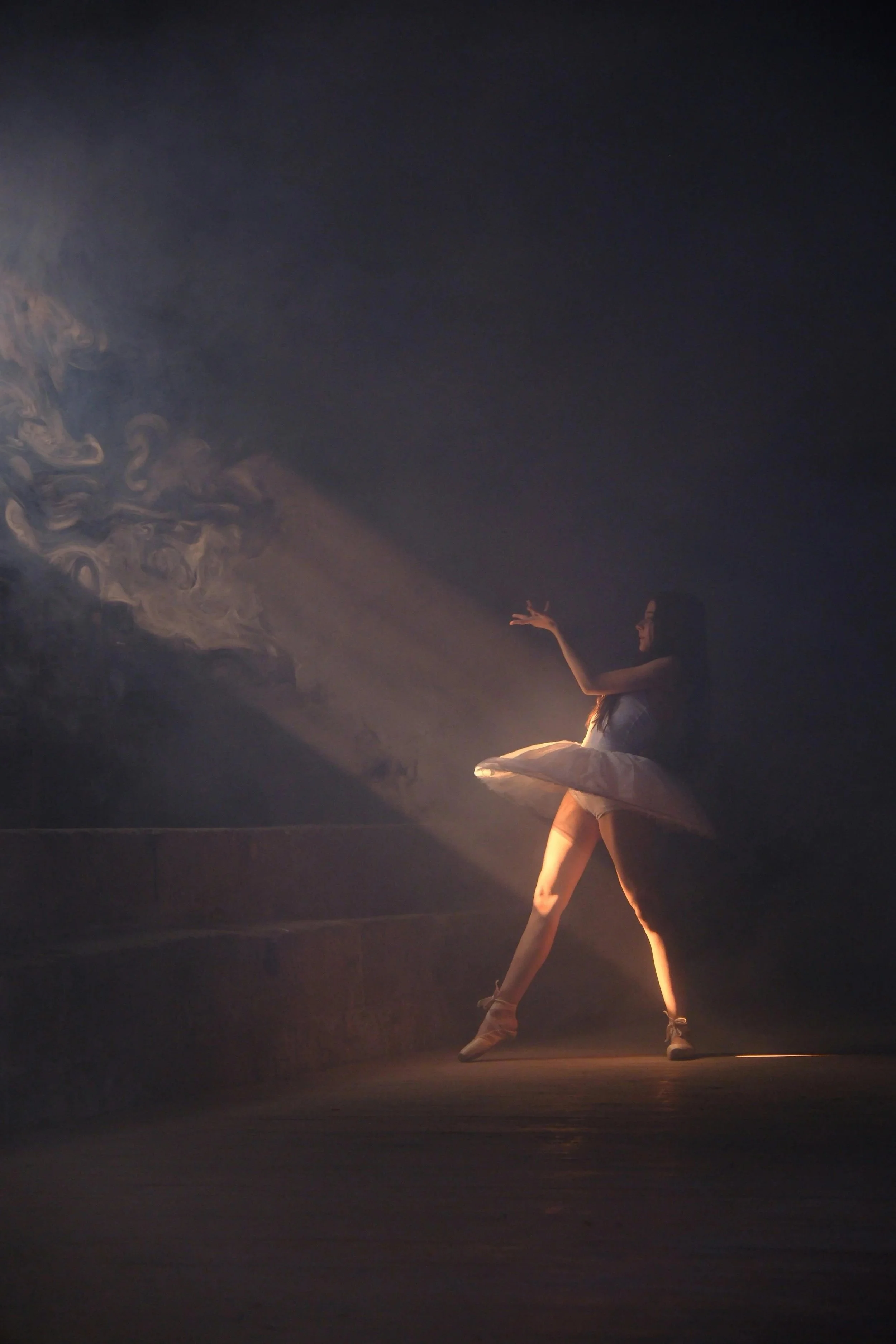 Bailarina en un escenario con iluminación suave, posando con un tutú y en medio de una danza, fondo oscuro.