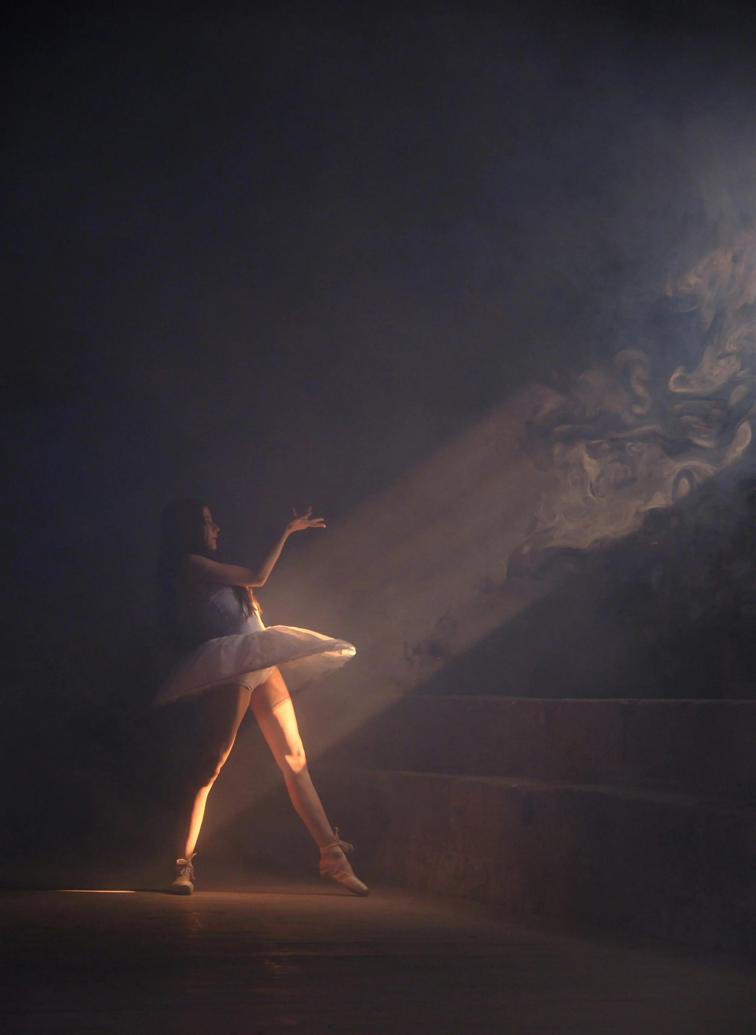 Bailarina en puntas con vestido de tutú rosa, en un escenario oscuro, iluminada por una luz suave que crea una atmósfera dramática.