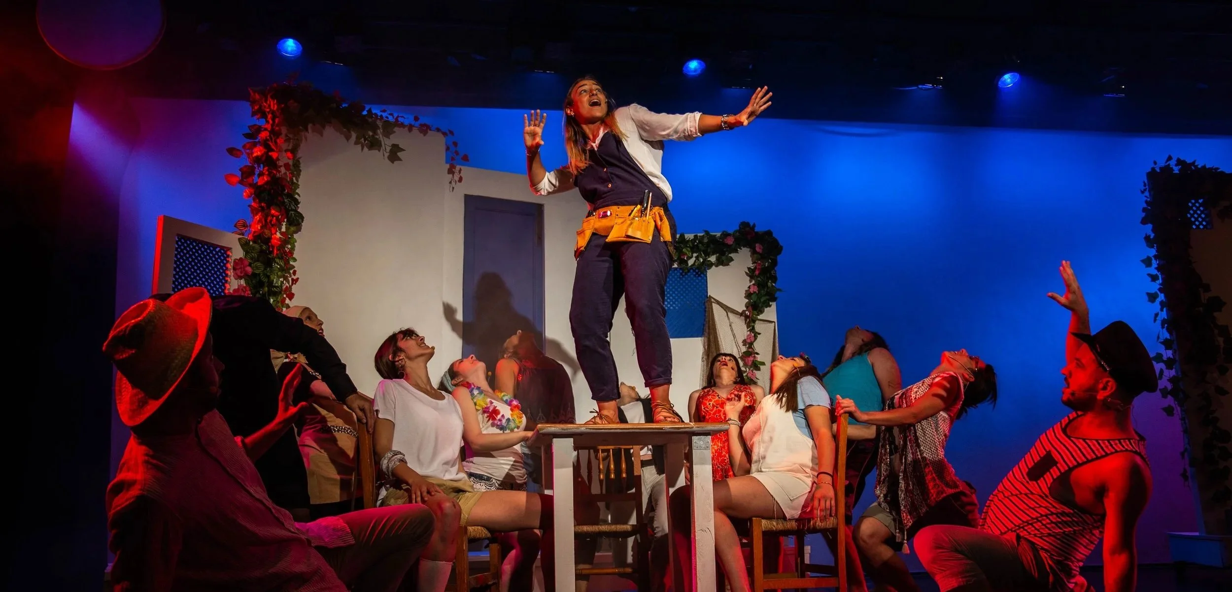 Escena de una obra de teatro con varias personas, una mujer en el centro está sobre una mesa levantando las manos, rodeada de otros actores en diversas posiciones y expresiones, decoraciones florales y fondos en tonos cálidos y azules.