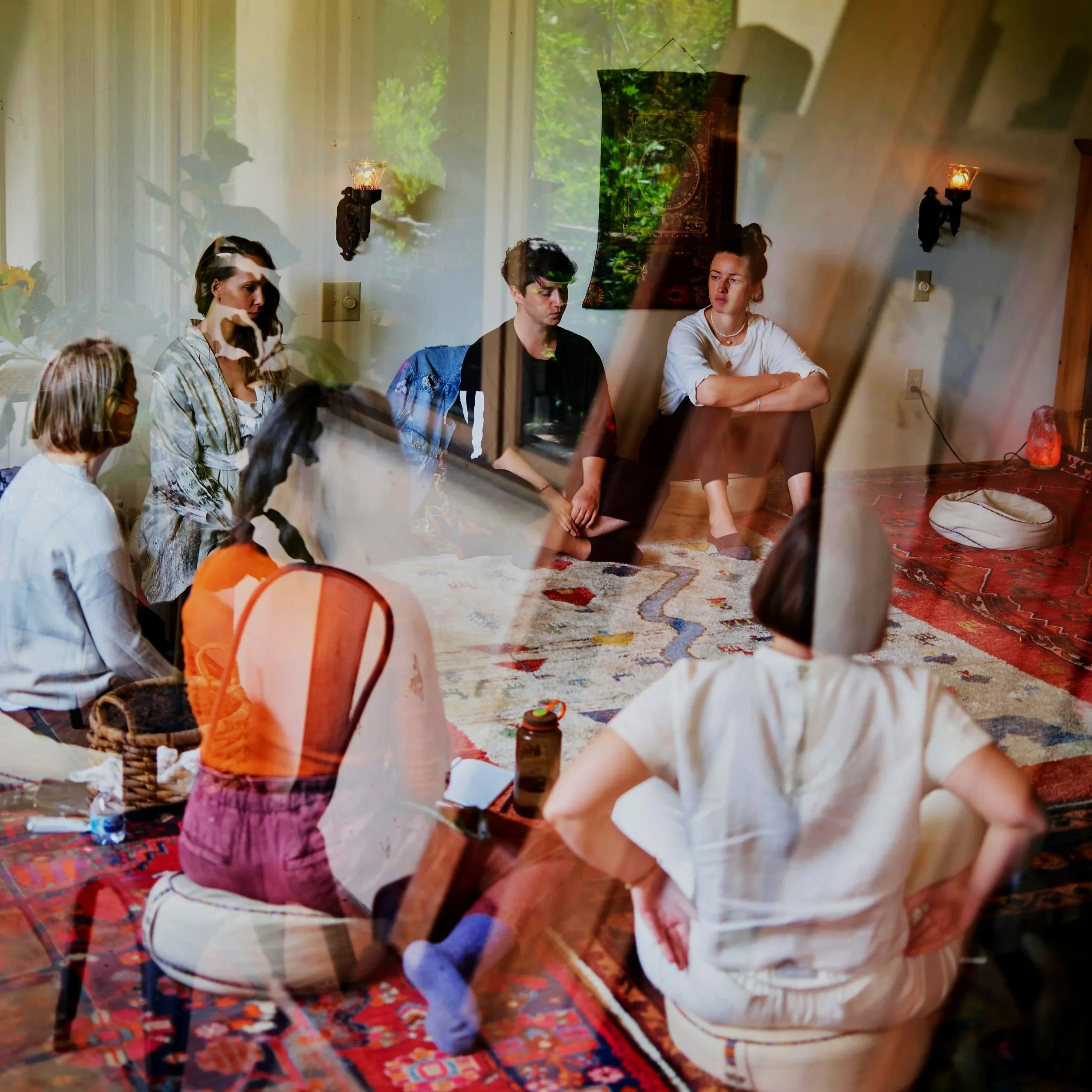 Grupo de personas en una habitación con alfombra, vistas desde afuera a través de una ventana, con plantas y paredes blancas de fondo.