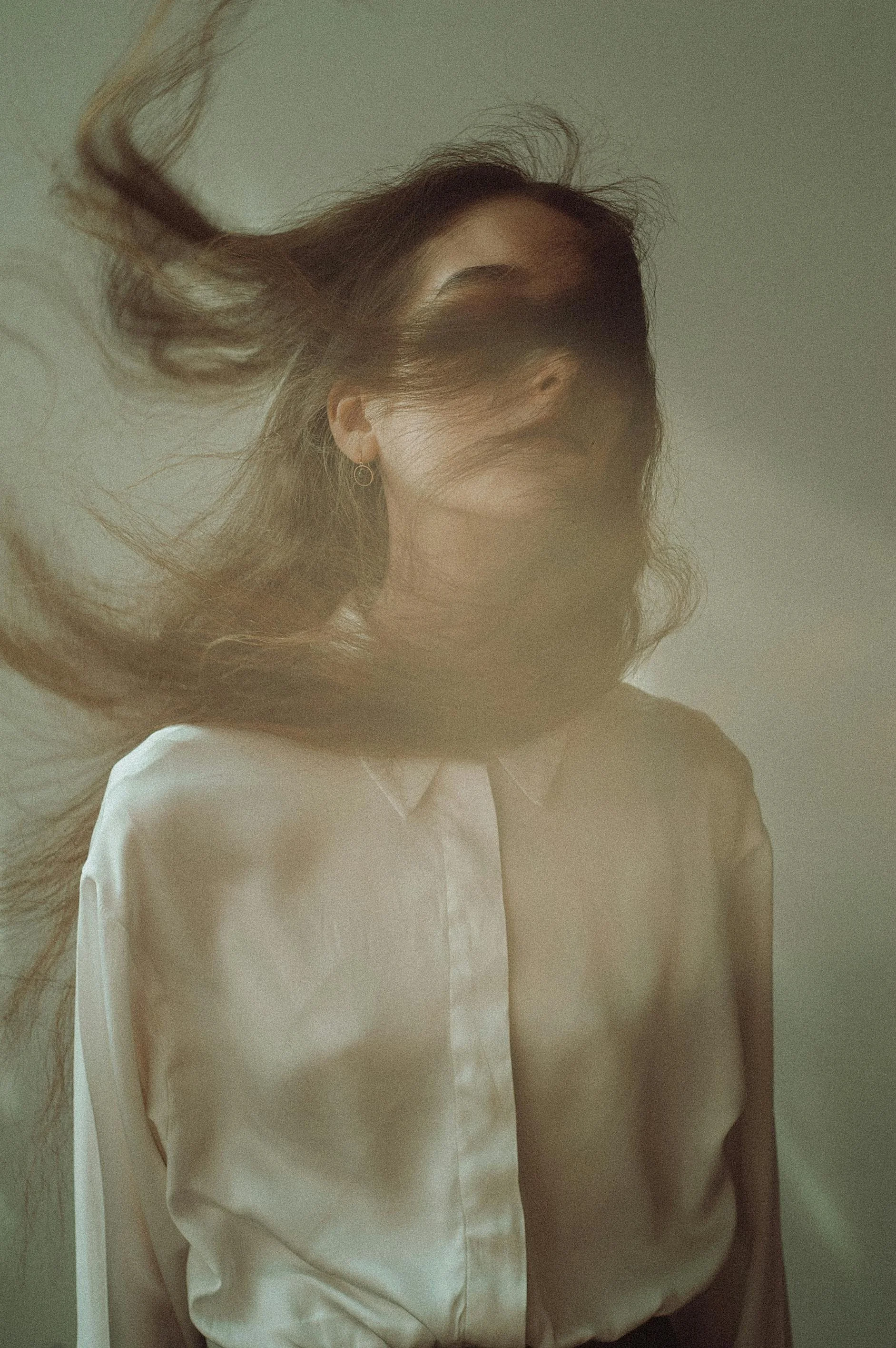 Una mujer con cabello suelto, de piel clara y usa una camisa blanca, en un fondo neutro, parece que está en movimiento o en una expresión artística.