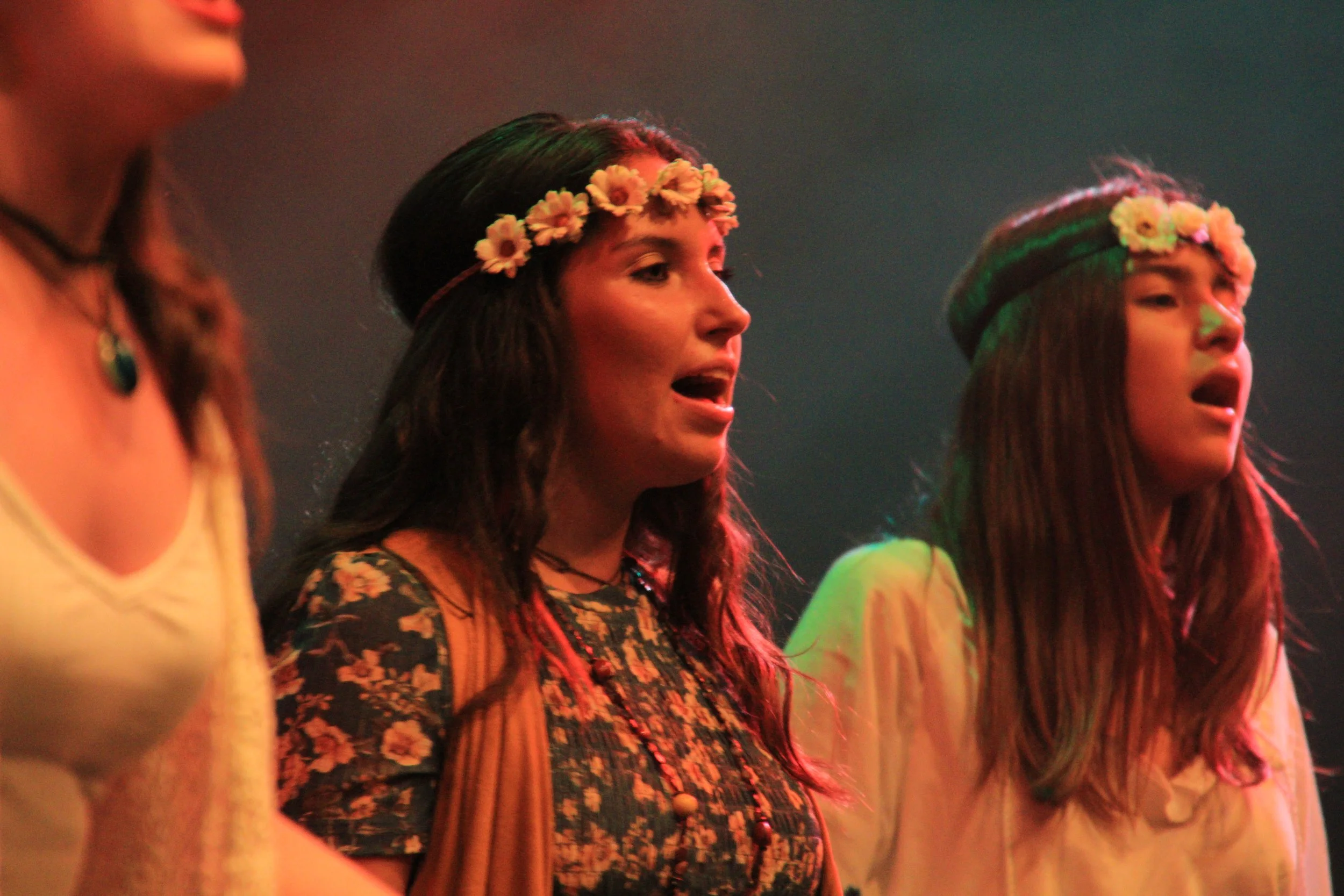 Tres jóvenes cantando en un escenario, usando coronas de flores en sus cabezas.