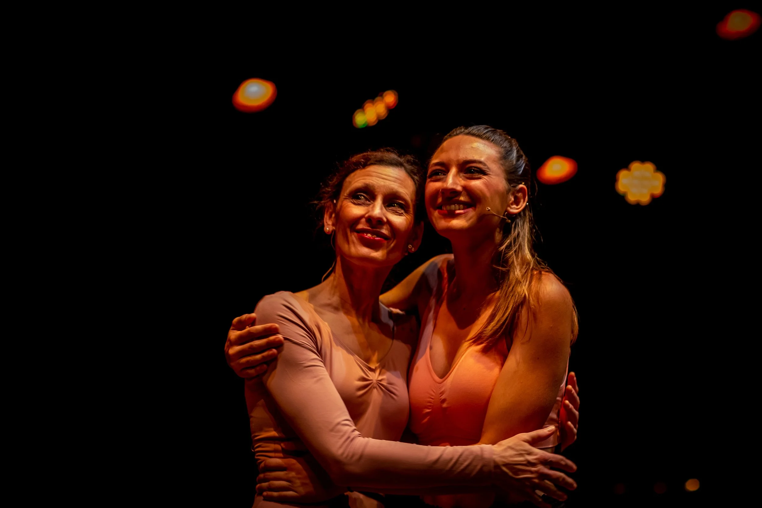Dos mujeres abrazadas, sonriendo en un escenario con luces difusas en el fondo, durante una presentación o celebración.
