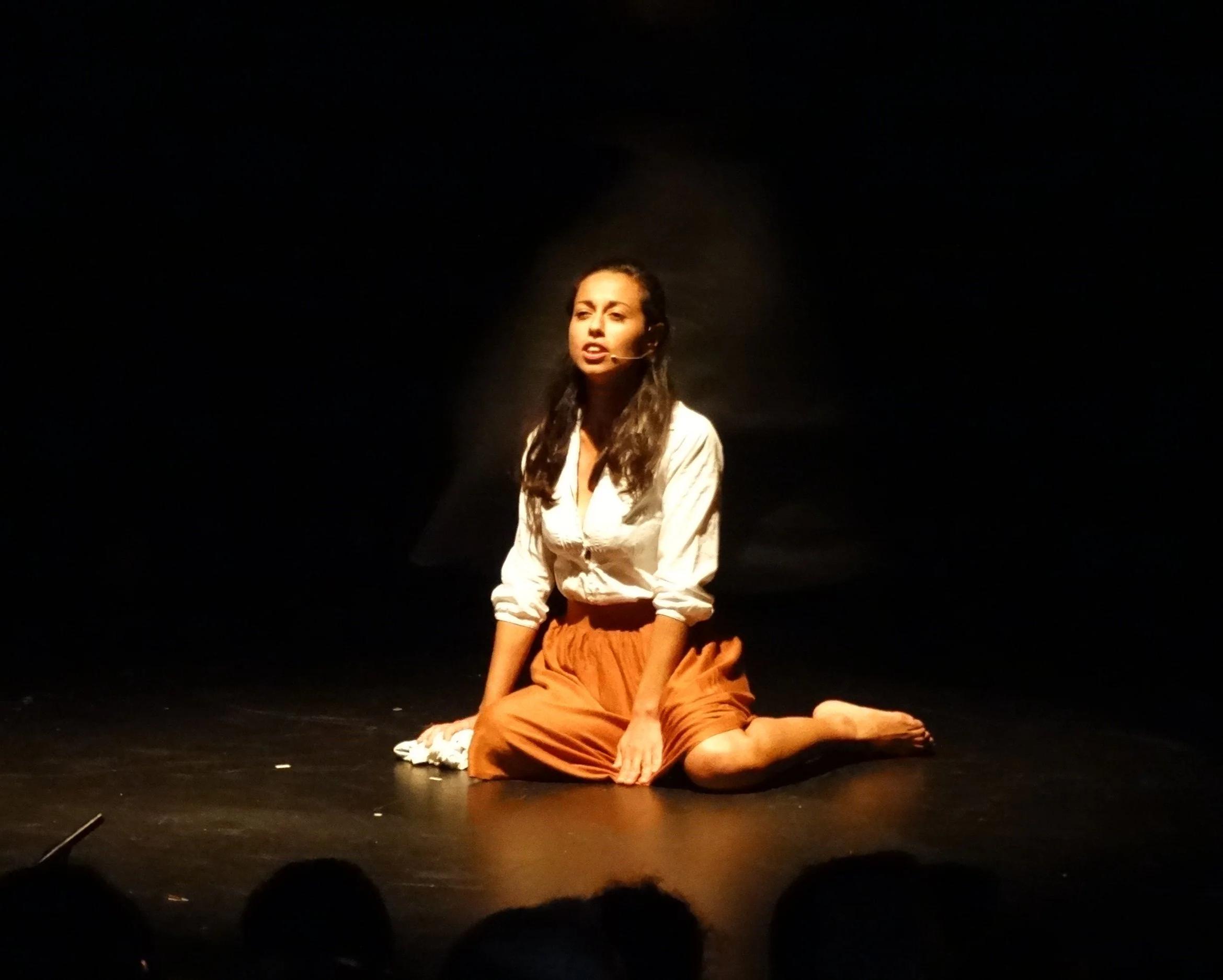 Una mujer participa en una obra de teatro, está en cuclillas en el escenario con un micrófono y expresión emocional, vestida con blusa blanca y falda larga color ocre, en un fondo oscuro.