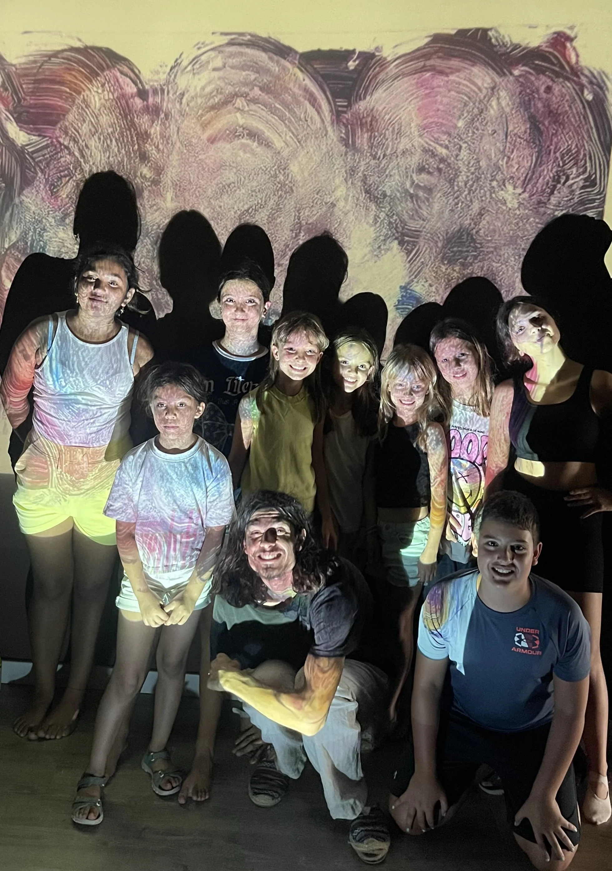 Grupo de niños y un adulto posando frente a una pared con proyección de arte abstracto, algunos niños están sonriendo, algunos siendo iluminados por luz y proyectado en la pared.