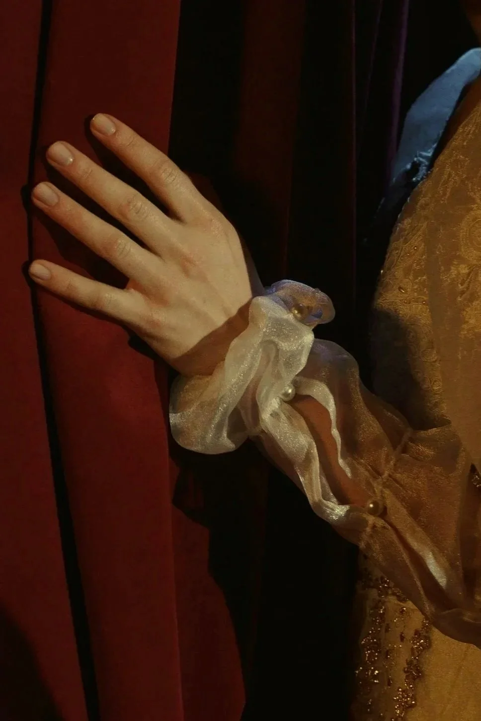 Una mano con un puño de tela dorada, adornada con perlas, en una pose sobre una superficie roja, con ropa elegantemente confeccionada.