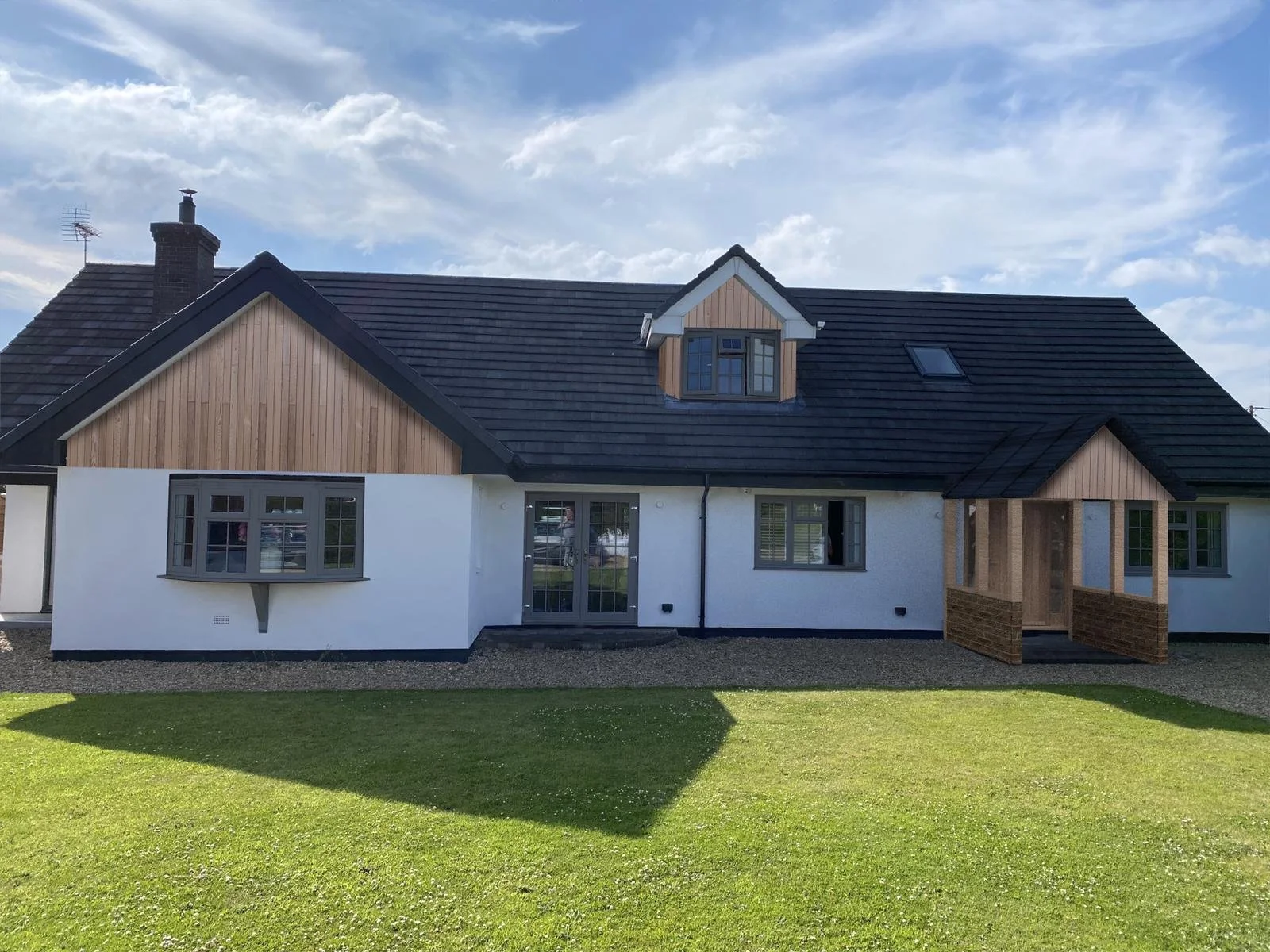 A modern house with white walls, wooden accents, a dark pitched roof, and a small porch, surrounded by a green lawn under a partly cloudy sky.