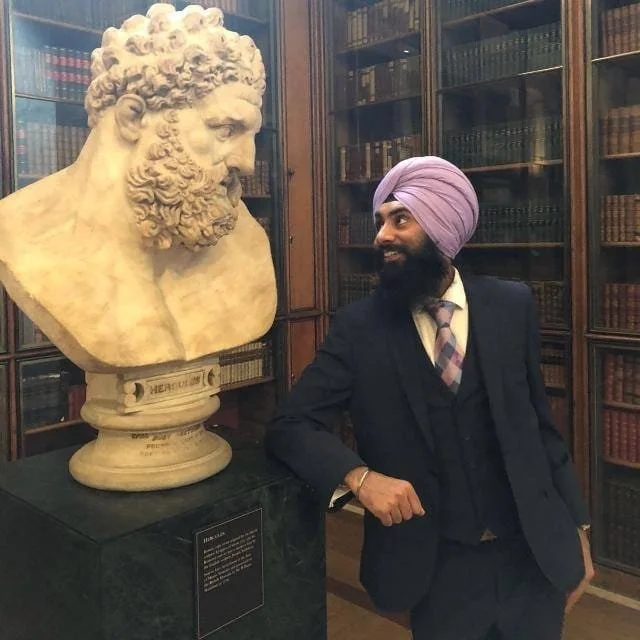 A man wearing a pink turban and a suit standing next to a large marble bust of a bearded man in a library filled with books.