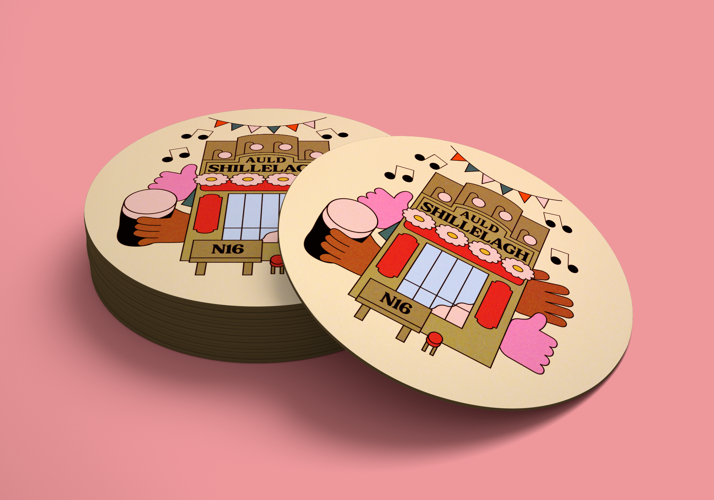 Close-up of circular coaster featuring a colorful illustration of a building with the words "Auld Shillelagh" on it, decorated with musical notes and pink flowers, set against a pink background.