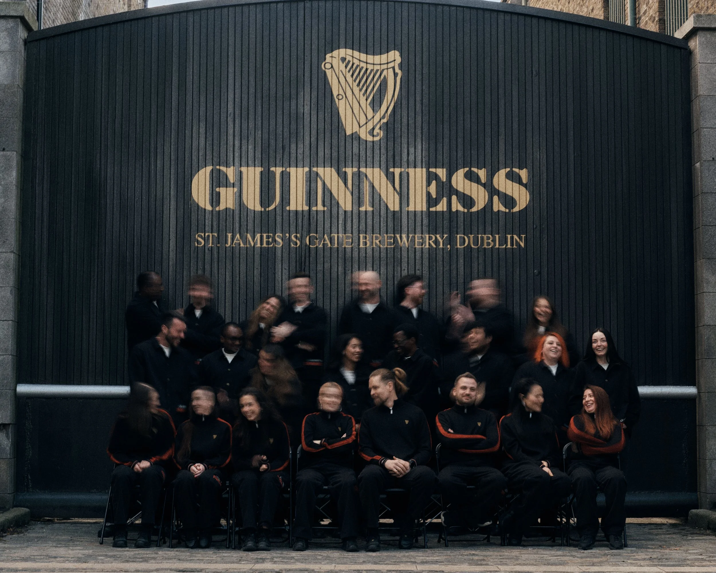 Group of people in black uniforms sitting and standing in front of a large Guinness sign at St. James's Gate Brewery in Dublin, with some individuals smiling and others blurred due to motion.