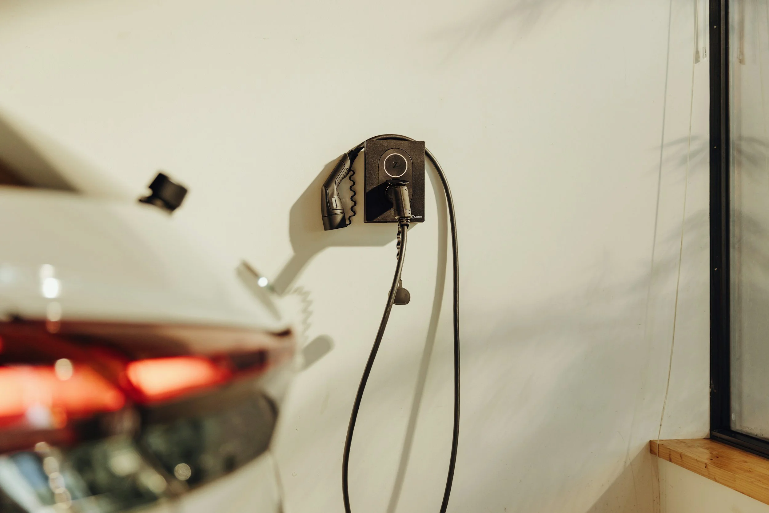 Electric vehicle charging station on a white wall with a black charging cable connected, partially visible car in the foreground.