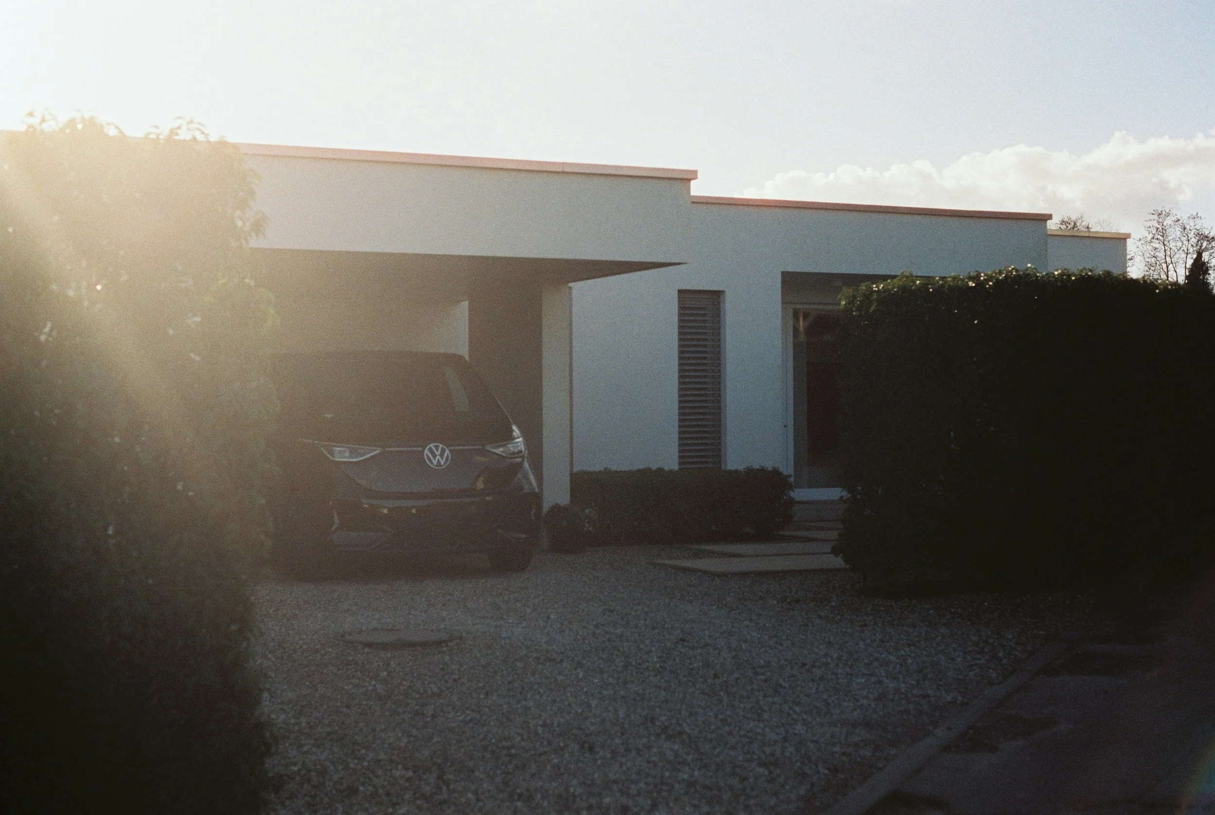 Modern house with white exterior, carport, Volkswagen EV electric car parked outside, surrounded by greenery, during sunset.