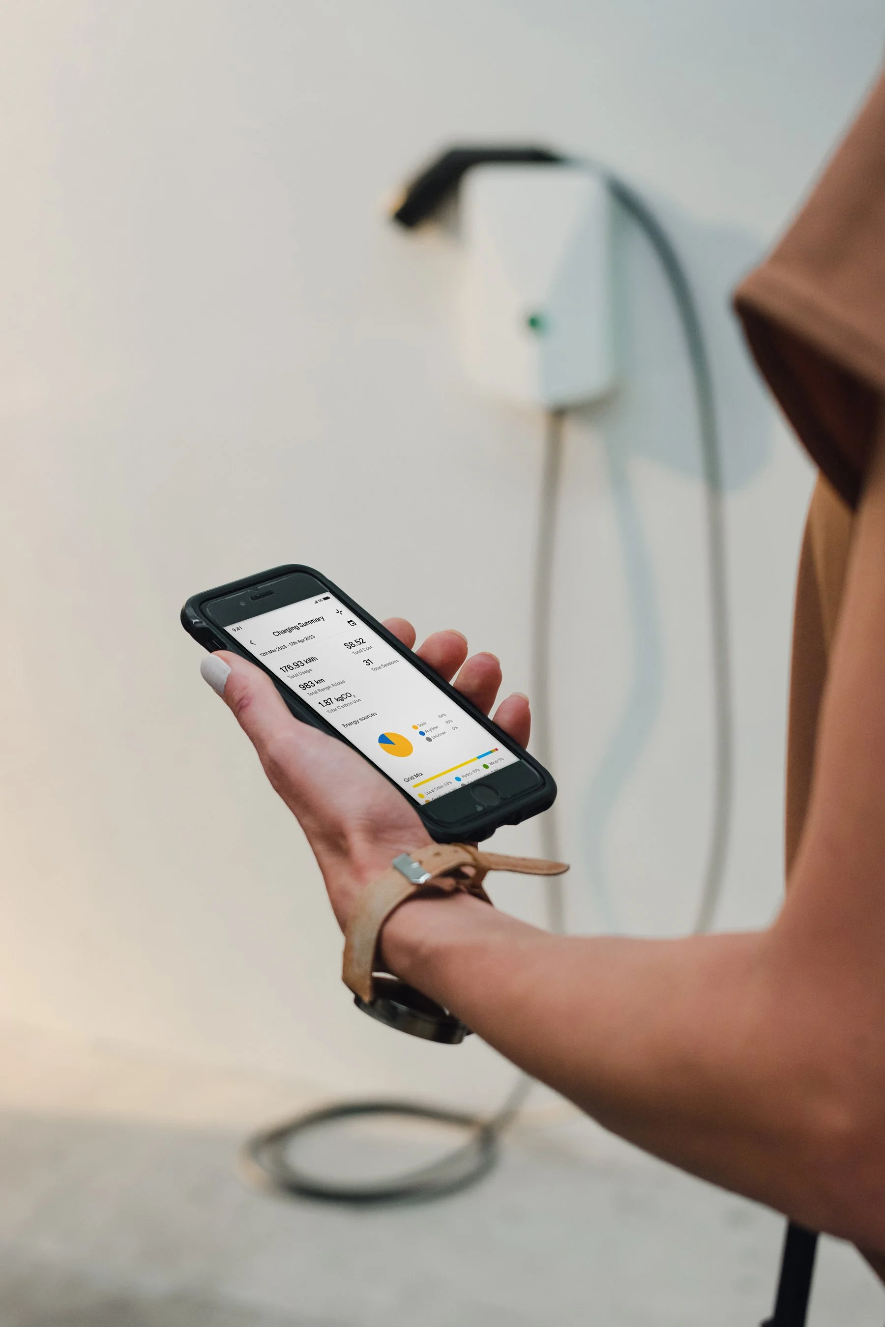 Woman holding iPhone, looking at charging summary displayed within the Evnex app, with Evnex E2 Plus smart charger installed on wall in background