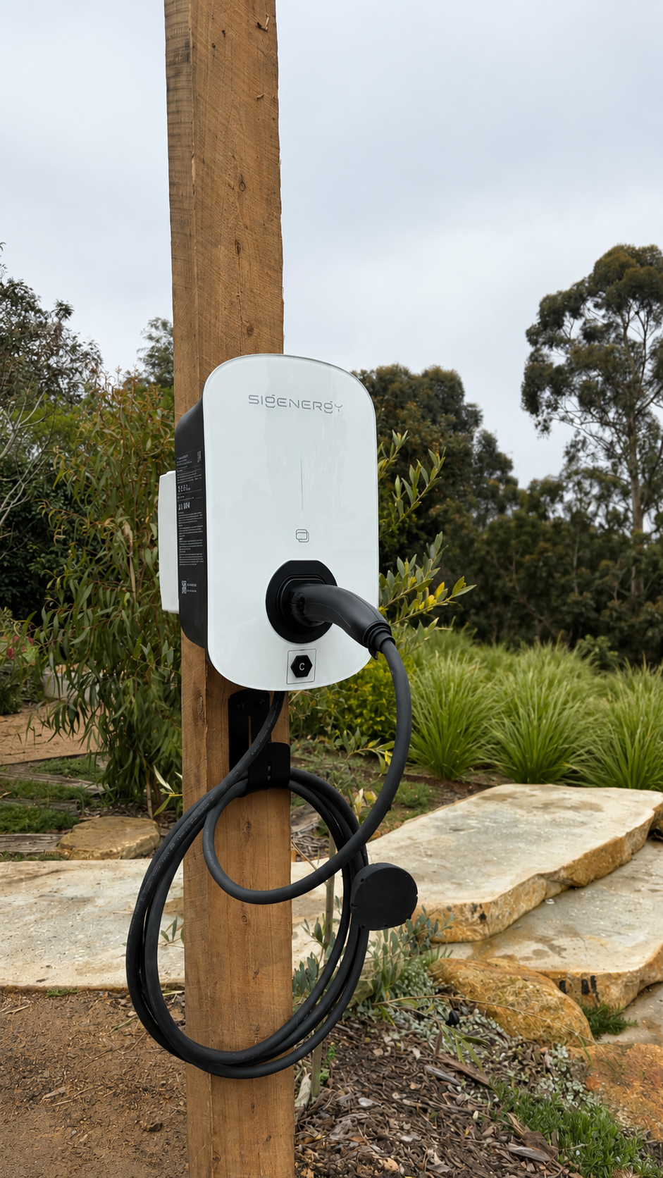A Sigenergy EV charger mounted on a wooden post in a leafy Greensborough garden area with rocks and bushes, cloudy sky in the background.