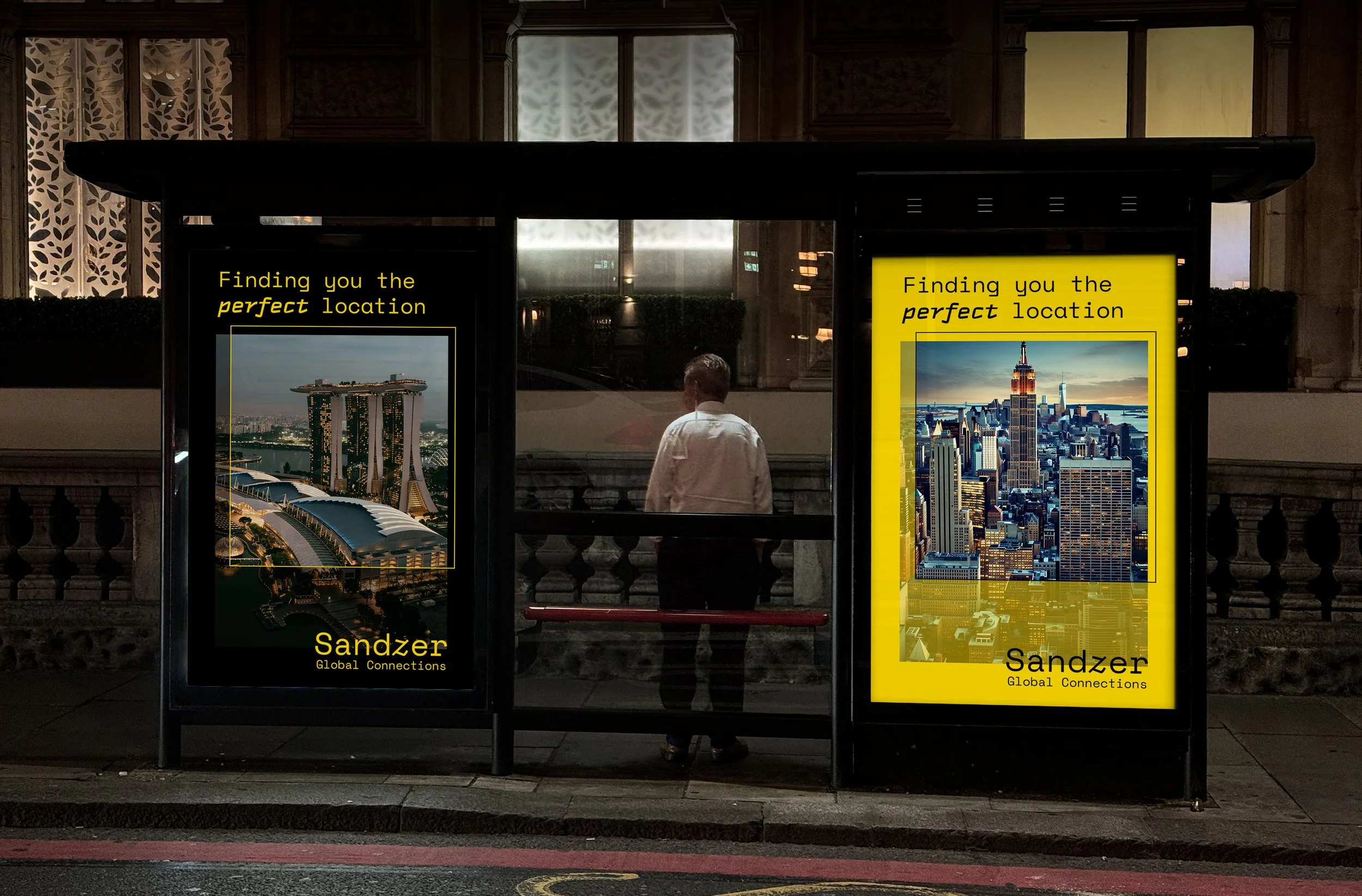 A bus stop with two advertisement posters featuring cityscapes, with a man standing in between, viewed from behind at dusk.