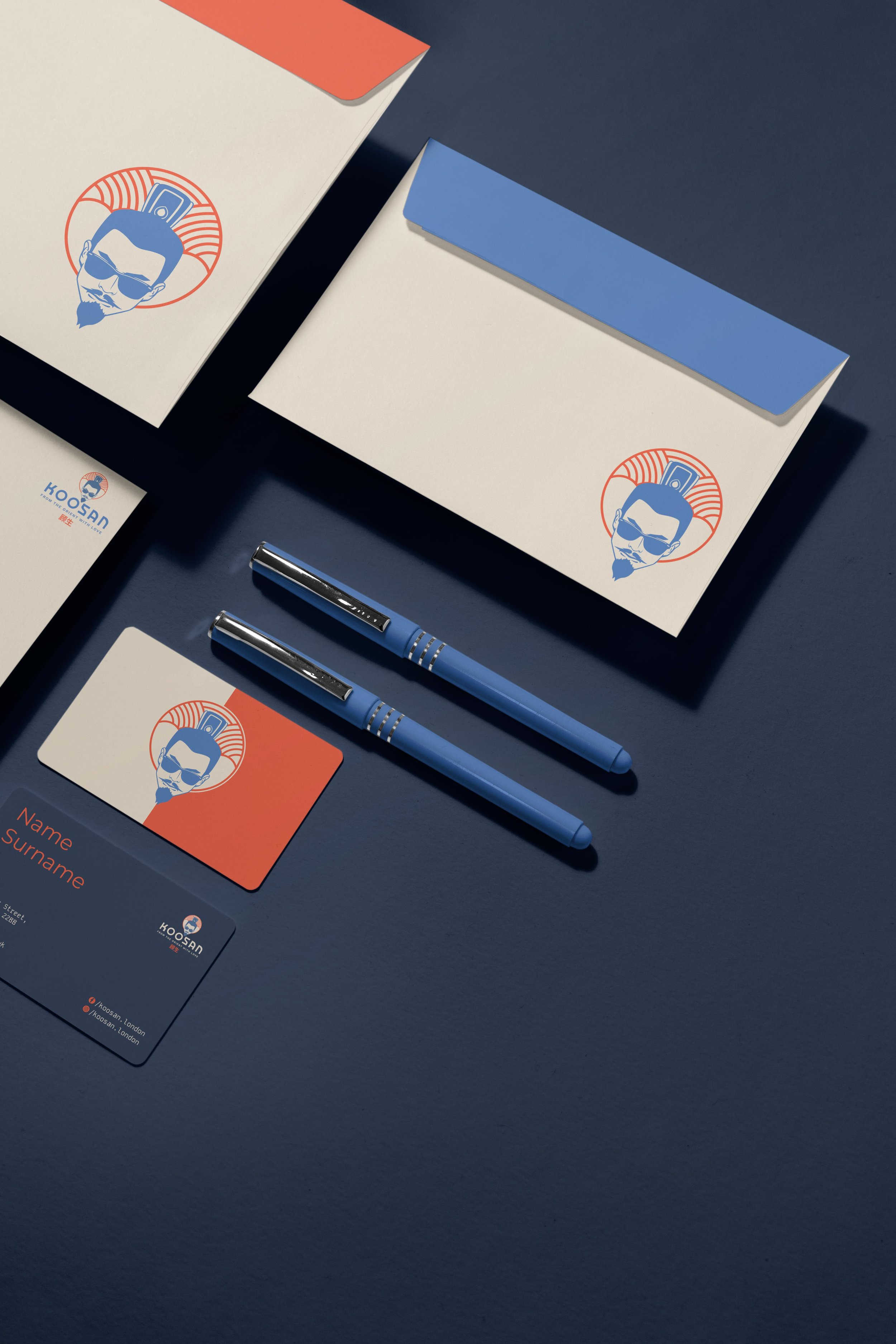 Stationery set including two blue pens, two white envelopes with blue and orange flaps, a business card, and an envelope, all featuring a logo of a man's face with sunglasses and a goatee, on a dark blue surface.