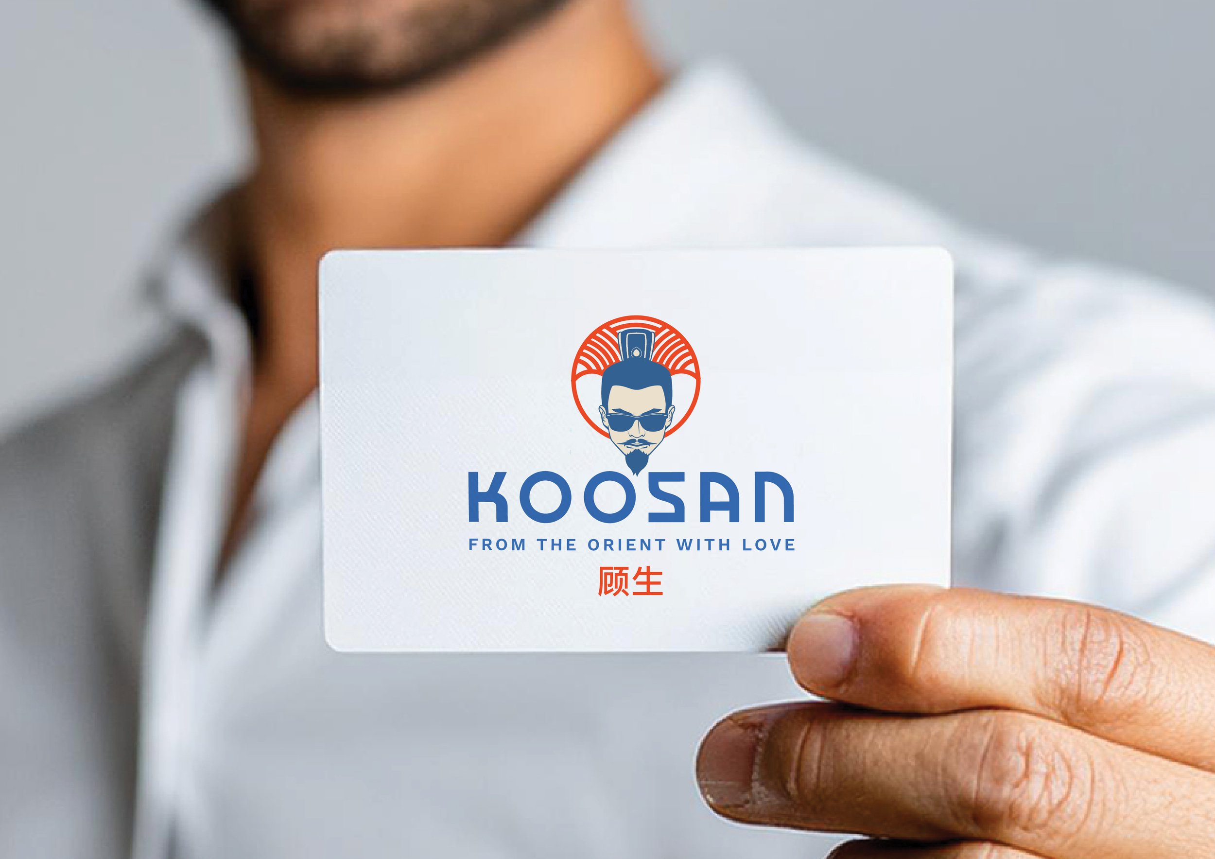 Person holding a white card with a logo of a stylized man with sunglasses and topknot hairstyle, accompanied by the words 'KOOSAN' and the tagline 'FROM THE ORIENT WITH LOVE' in blue text, and red Chinese characters beneath.