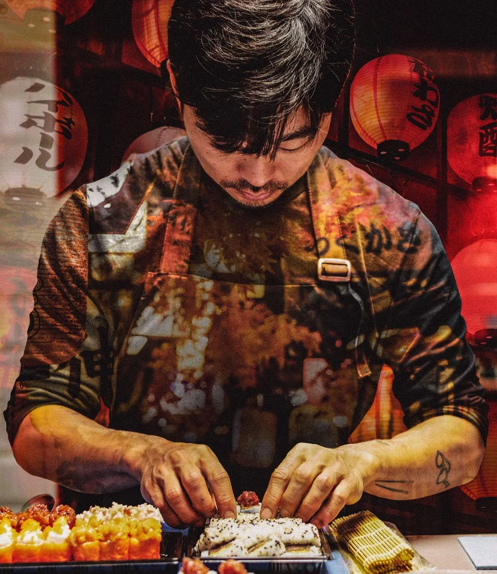 A man preparing sushi at a sushi bar with Japanese lanterns in the background.