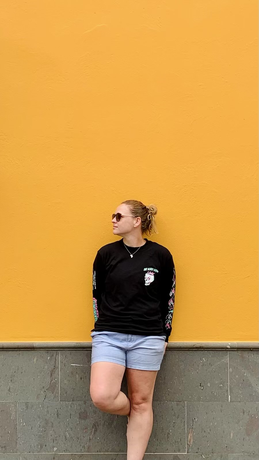 A woman leaning against a yellow wall, wearing sunglasses, a black graphic long-sleeve shirt, and light blue shorts.