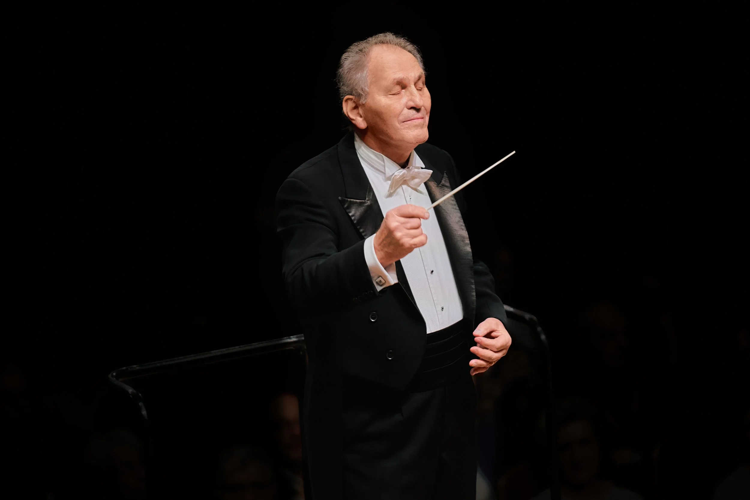 Older man in formal tuxedo conducting an orchestra, holding a baton with closed eyes and a focused expression, against a black background.