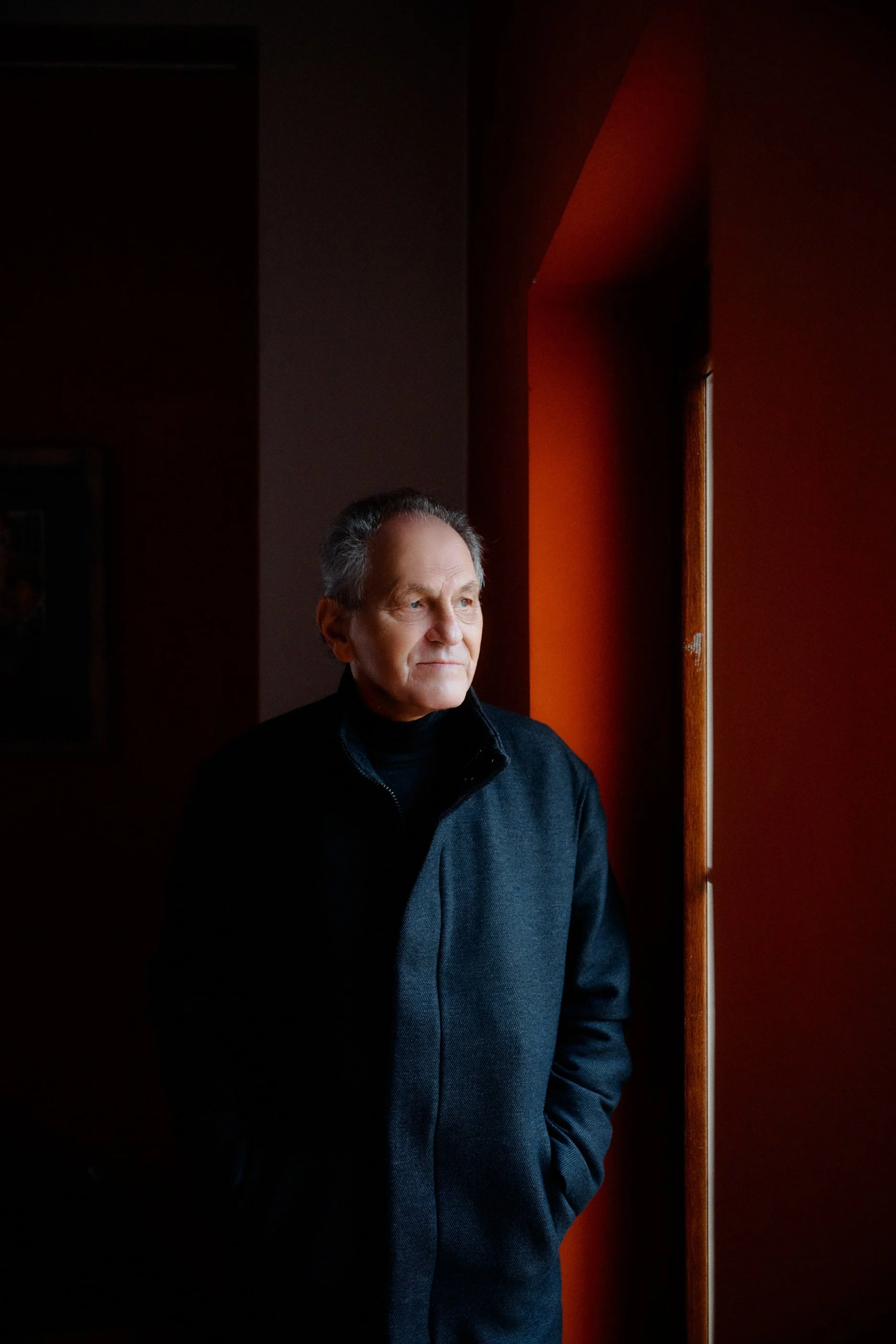 Older man with gray hair dressed in black coat standing near a window in a dimly lit room with red walls, looking out thoughtfully.