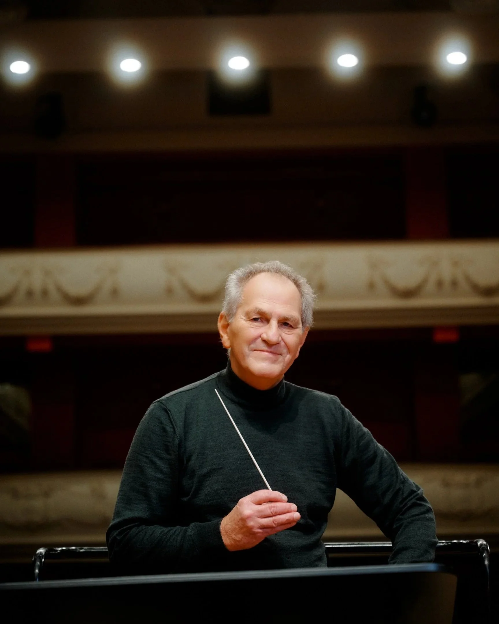 An older man with gray hair wearing a black turtleneck sweater stands on a stage and holds a conductor's baton in his right hand.