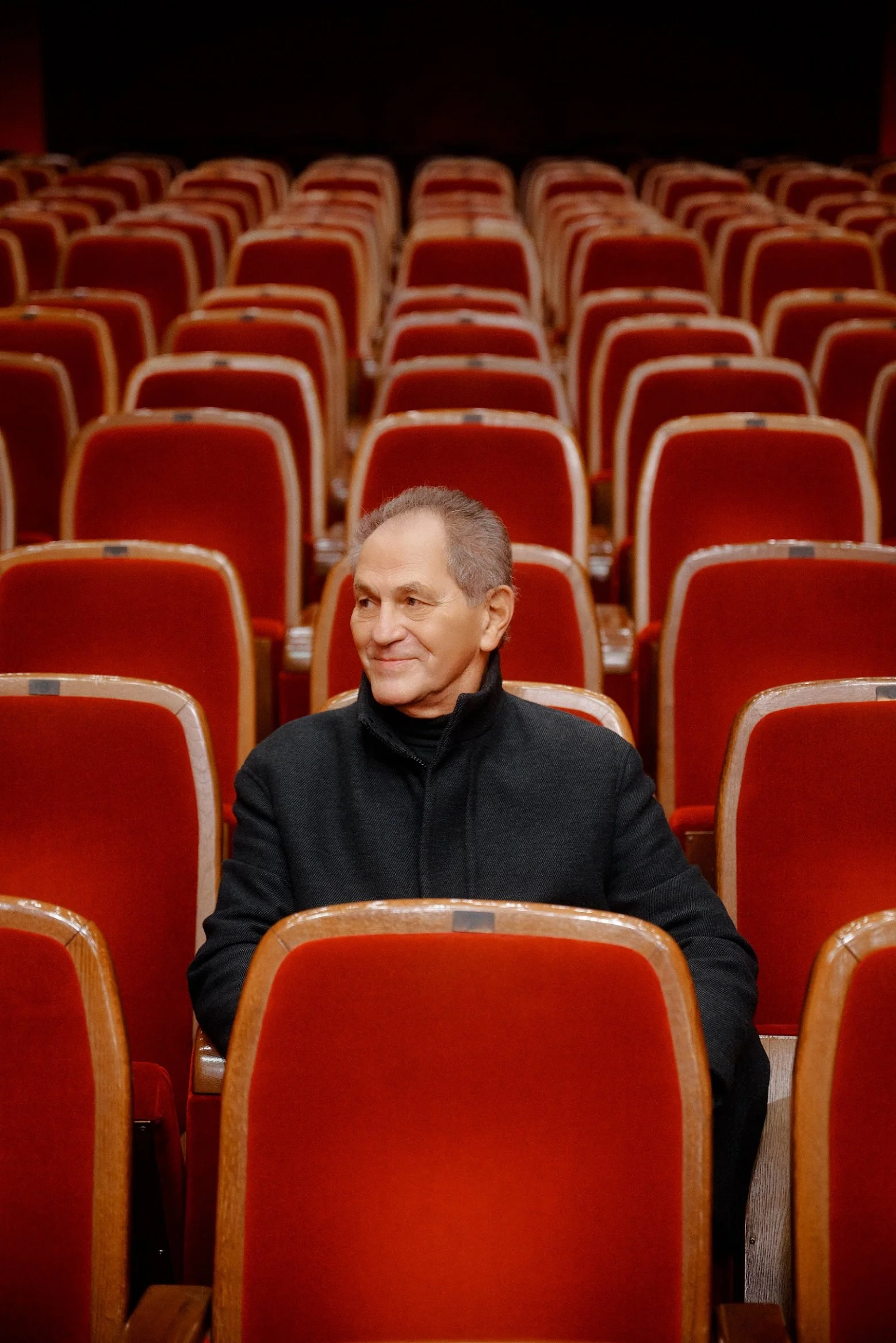 A man sitting alone in red theater seats, smiling, in an empty theater.
