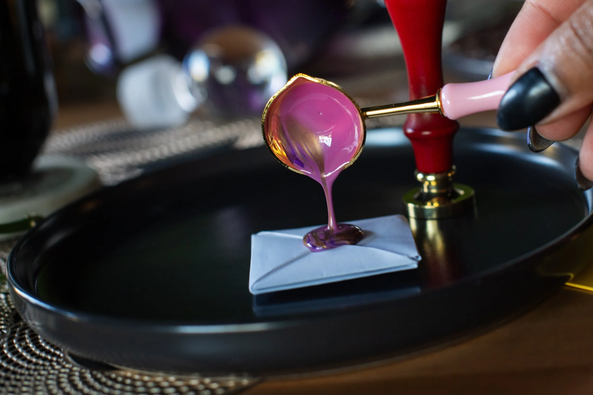 A person holding a pink and gold jewelry loupe over a pink gemstone. The gemstone is resting on a white paper on a black tray, with blurred objects in the background.