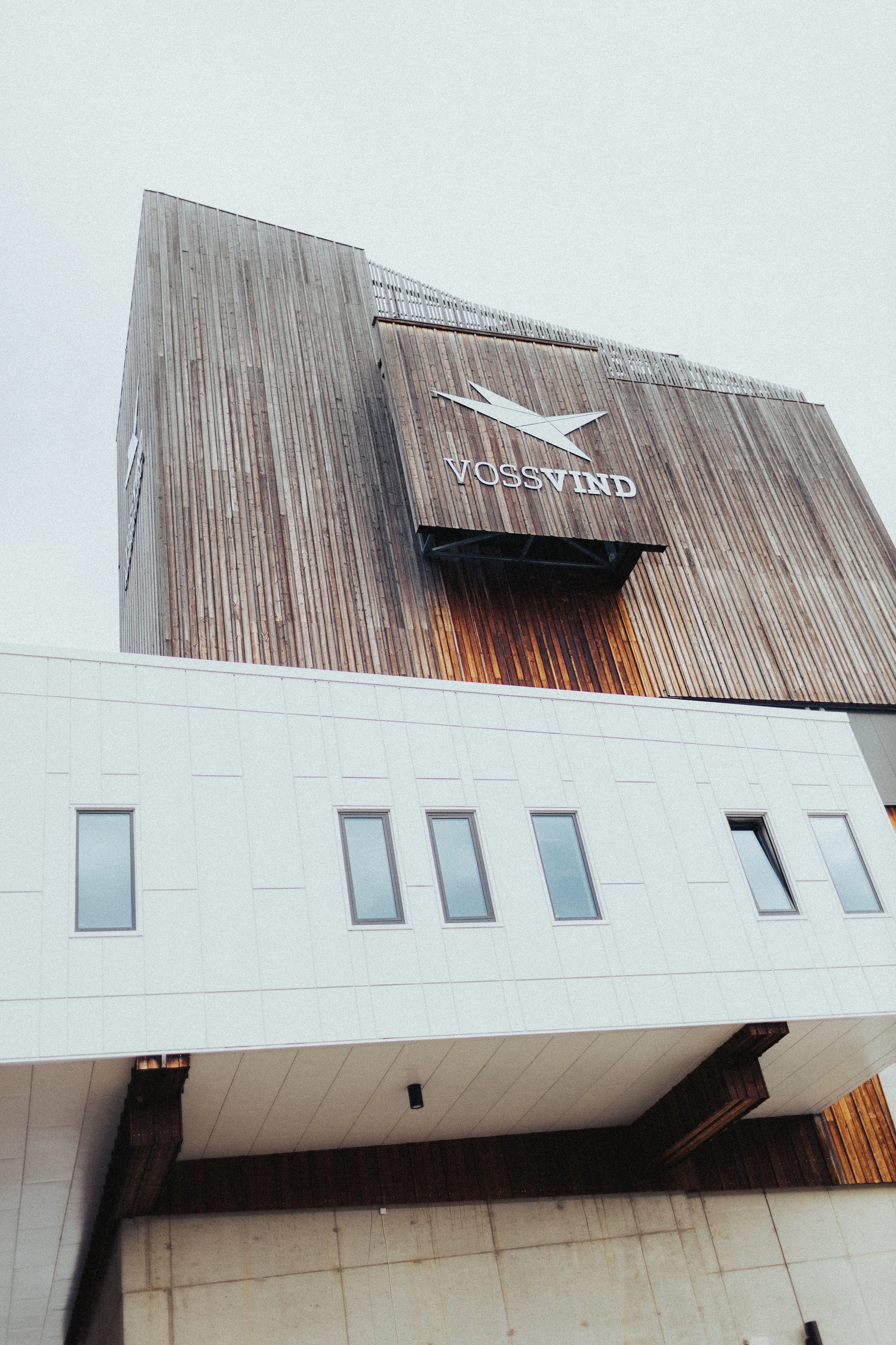 Bilde av et moderne høyt bygg, overflater av treverk og hvite paneler, med en stor VossVind-logo på fasaden.
