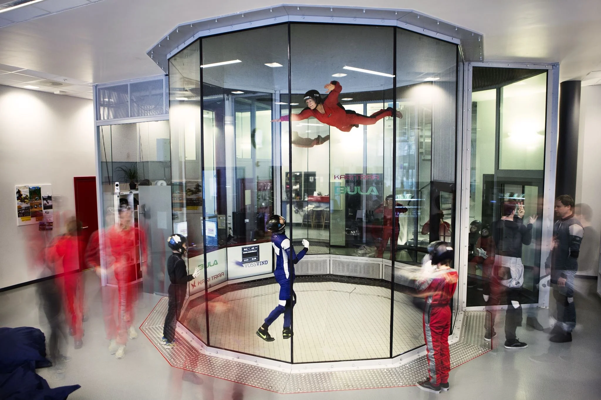 Innendørs skydiving simulator med flere personer, noen venter i kø, noen flyr, og andre observerer.